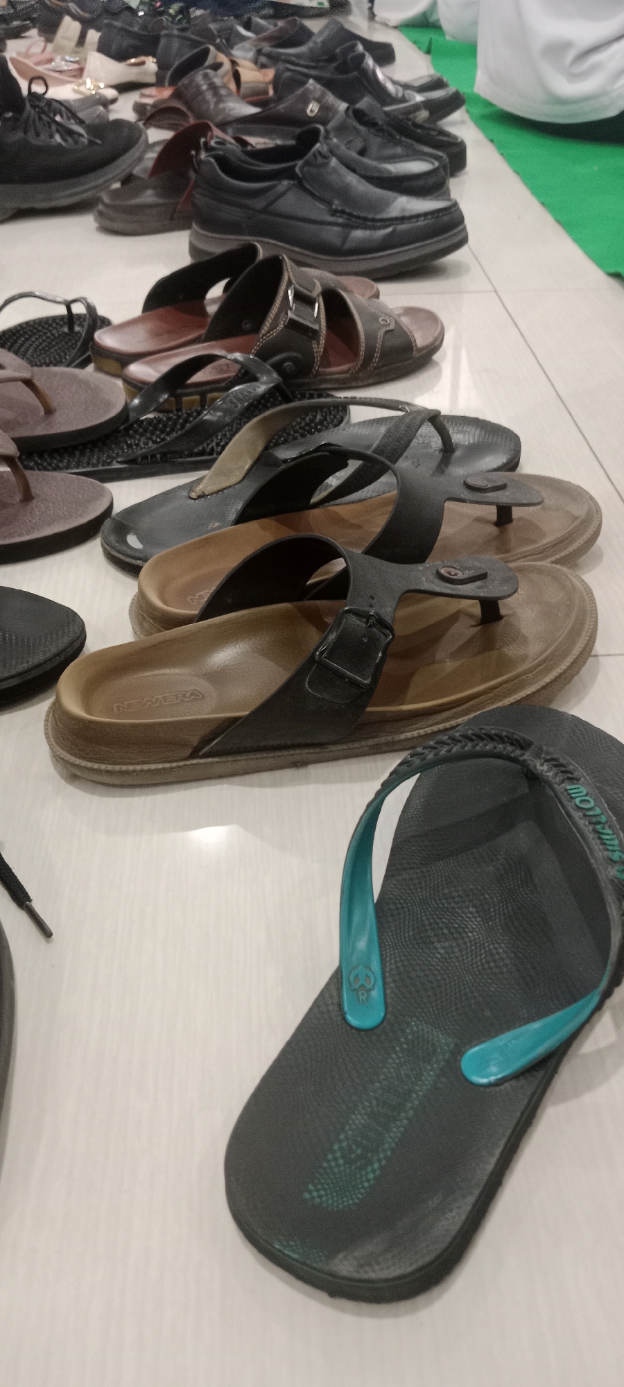 A collection of men's footwear, including various styles of sandals and shoes, are neatly arranged on a light-colored tiled floor, likely in a domestic or communal setting such as a mosque or a waiting area, in Tuban, Indonesia. The footwear varies in color and material, with some appearing to be leather and others rubber or synthetic. The arrangement suggests they are either being stored or are waiting to be worn. The background shows a hint of green fabric, possibly a prayer mat or carpet, and other shoes blurred in the distance. The lighting appears to be artificial, casting subtle shadows.