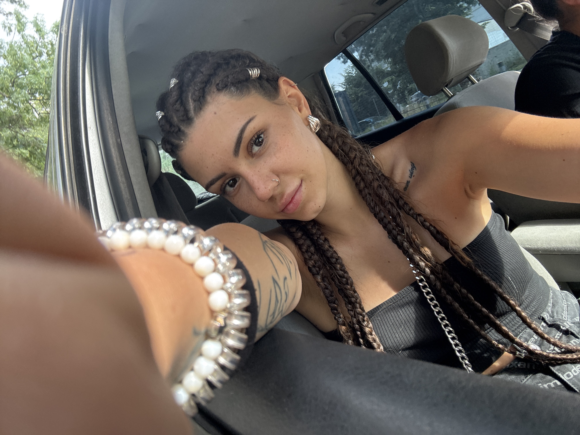 This is a selfie taken by a young woman inside a car. She has braided hair with metal rings woven into some of the braids. She is wearing a black tank top with a silver chain strap and a bracelet made of white beads and silver accents on her left wrist. A tattoo is visible on her left arm. She is wearing large, sculptural earrings and a nose ring.
The woman is looking towards the camera with a slight smile. The interior of the car is visible, including the headrest of the seat behind her. Outside the car window, trees and a building can be seen, suggesting an outdoor setting during the daytime. The lighting indicates it is likely daytime.
The scene appears to be casual, and the woman's expression suggests a relaxed or happy mood. There is no visible text in the image. The location context is Sofia, Bulgaria.