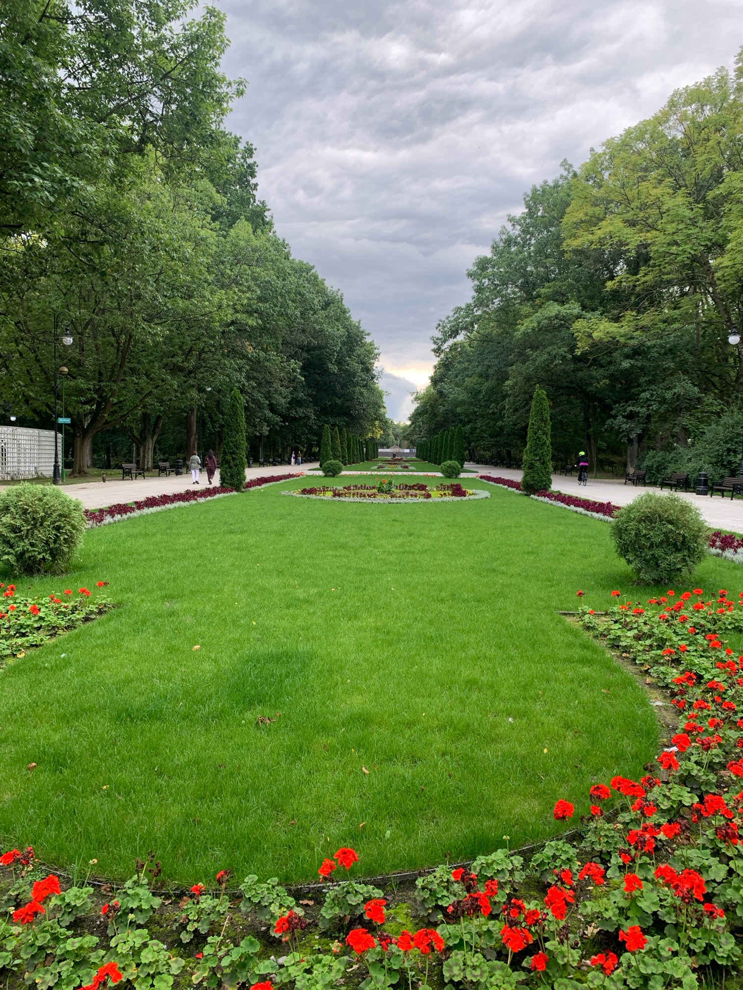 A vertical shot captures a manicured park with a central lawn bordered by flowerbeds and trees. The park features a winding pathway on either side of the central lawn. Along the pathway, benches are visible, and a few individuals are strolling or cycling. The sky is overcast with a mix of grey and white clouds. The scene is likely located in Branicki Park in Białystok, Poland, as indicated by the characteristic landscaping and architecture.
