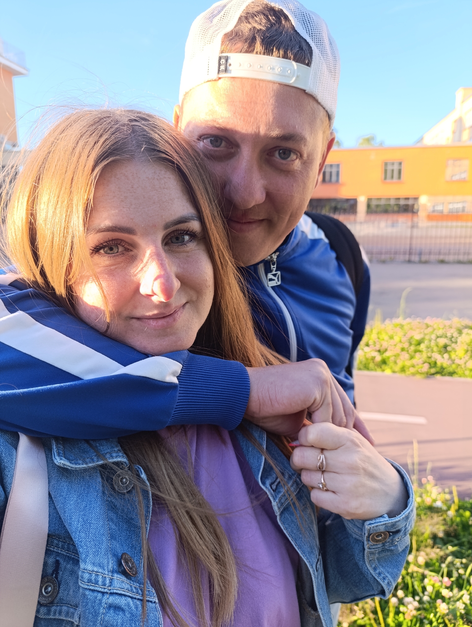 This is a selfie of a man and a woman, likely a couple, standing close together outdoors. The man, wearing a white baseball cap and a blue athletic jacket, has his arm around the woman. She is leaning into him, resting her head on his arm. She has long, auburn hair and is wearing a denim jacket over a purple top. Both appear to be smiling and looking towards the camera.
The background suggests an urban or suburban setting, with a bright orange building visible behind them. There is a paved area in the foreground, possibly a walkway or road, bordered by green grass and small white flowers. The sky is clear and blue, indicating a sunny day. The lighting suggests it is late afternoon or early evening, with the sun casting a warm glow on the woman's hair and face. The overall mood of the image is affectionate and happy.
There is no visible text in the image.