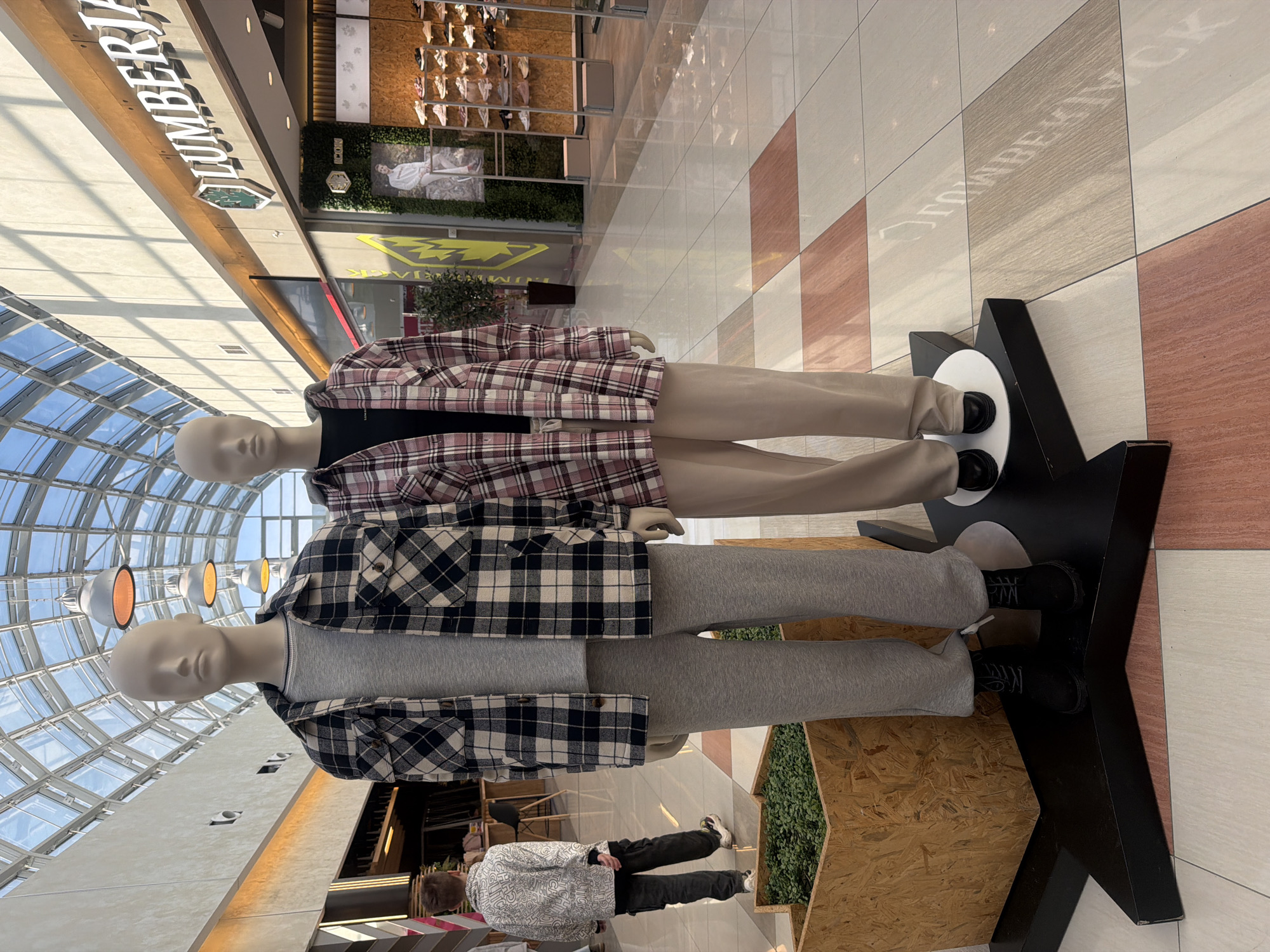 The image presents an interior view of a modern shopping mall, identified as the Planet Perm (ТРЦ "Планета") located at Shosse Kosmonavtov, 162Б, Perm, Russia. In the foreground, two headless mannequins are positioned on a black star-shaped display platform. The mannequin on the left, designed to represent a male, wears a grey long-sleeve t-shirt, an open black and white plaid flannel shirt with two chest pockets, grey sweatpants, and black lace-up boots. The mannequin on the right, designed to represent a female, wears a black t-shirt, an open pink and white plaid flannel shirt, beige wide-leg trousers, and black slip-on shoes.
In the mid-ground, a child, appearing male, is seen from behind, walking away to the left. He wears a white long-sleeved shirt with an abstract dark pattern, dark cargo-style pants, and sneakers with white soles.
The mall's environment features a high vaulted ceiling constructed of glass panes, allowing natural light to enter. Several large, metallic, dome-shaped pendant lights hang from the ceiling. The floor is tiled with large square tiles arranged in a checkerboard pattern, utilizing shades of light grey, white, and reddish-brown. To the right, the facade of a "LUMBERJACK" store is prominent, displaying its name in white lettering with a green tree logo. The store interior, visible through the entrance, showcases numerous pairs of shoes on wall-mounted shelves and a large poster featuring a person. Elements of artificial green foliage decorate the store's exterior wall. A wooden planter containing small green plants is situated behind the left mannequin. Reflections of ceiling structures and store names are visible on the polished floor tiles.