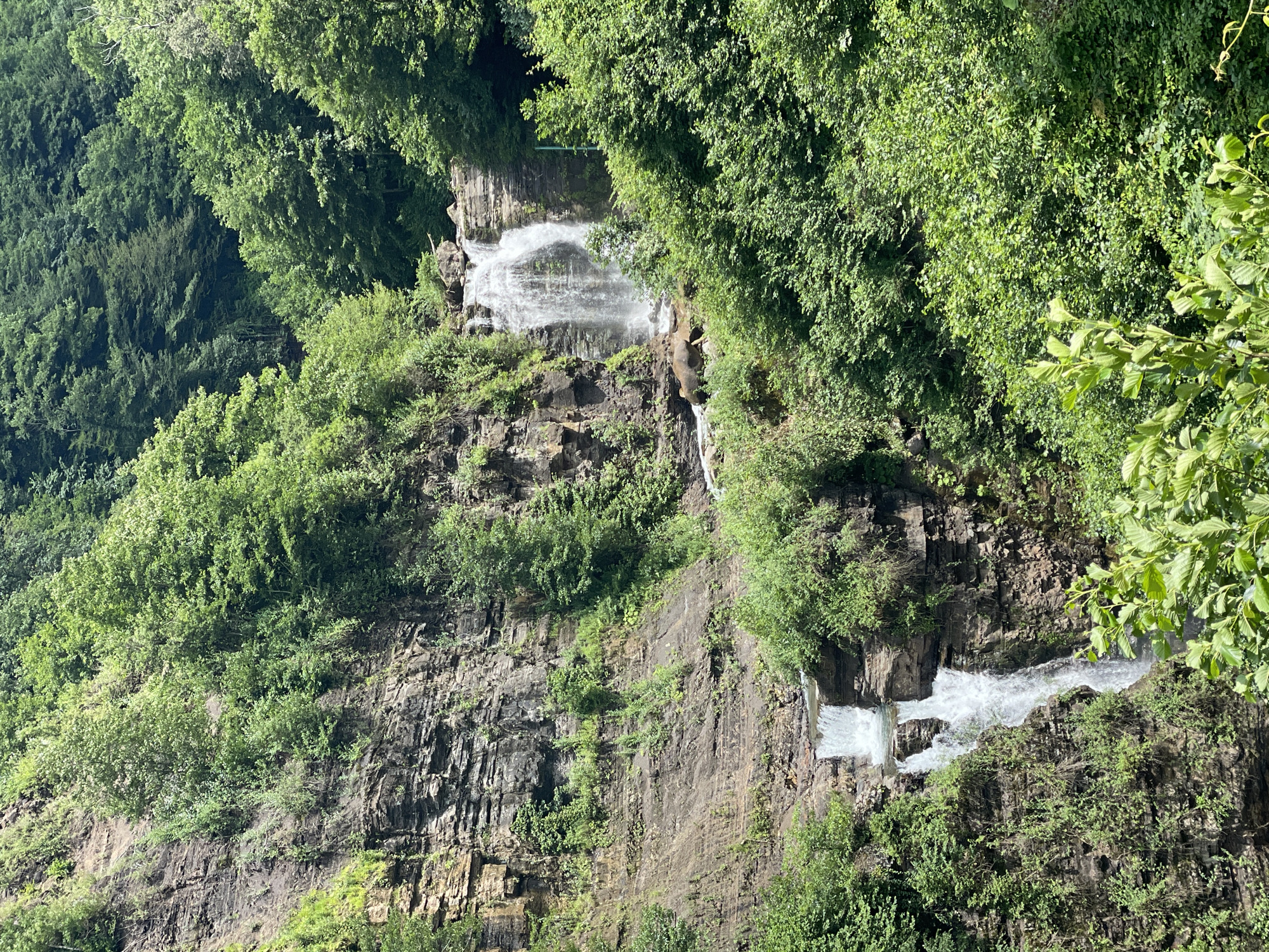 This image depicts a multi-tiered waterfall cascading down a rocky cliff face, surrounded by lush green vegetation and dense forest. The waterfall appears to be located in a natural, undeveloped area, consistent with a remote wilderness setting. The cliff is characterized by layered rock formations and is heavily overgrown with plants and trees. The water is white and foamy as it falls, creating a dynamic contrast with the static rock and foliage. The overall scene suggests a tranquil and picturesque natural landscape.