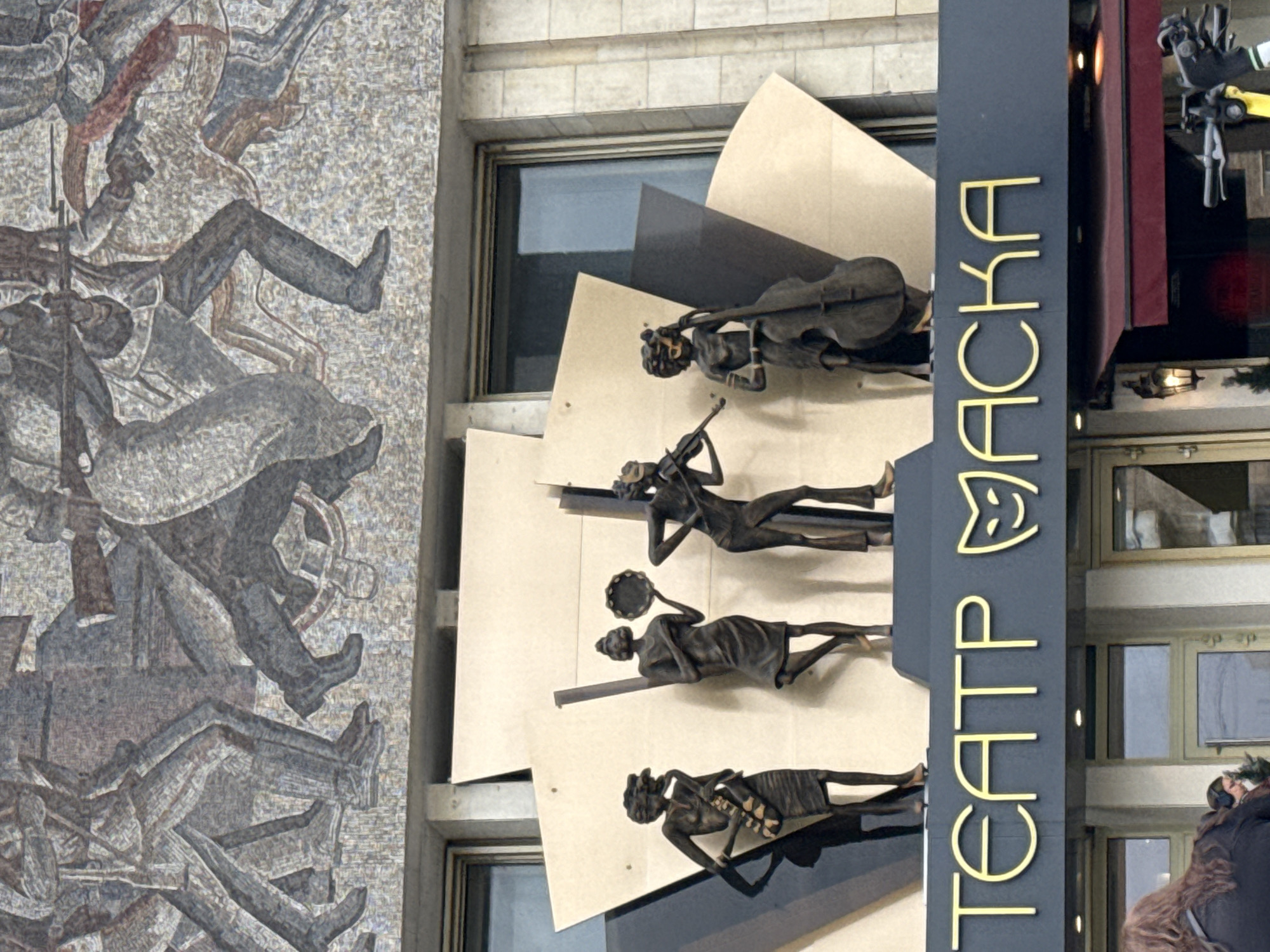 This vertical image captures a portion of a building facade in Moscow, Russia, presenting a juxtaposition of historical art and modern theatrical advertising.
On the left, a large mosaic mural dominates the scene. It depicts numerous figures, likely soldiers or revolutionaries, in a dynamic and crowded composition, some holding what appear to be rifles or weapons. The style is fragmented and textured, suggesting a scene of struggle or historical significance.
Adjacent to the mosaic, on the right side of the building, is a tall, dark vertical sign with prominent gold lettering. The text reads "ТЕАТР МАСКА" (TEATR MASKA), and above the word "МАСКА", there is a stylized mask icon, characteristic of theatrical venues. Adorning the facade around this sign are several bronze-colored, stylized figures of musicians. Visible instruments include a cello, a violin, and a tambourine, further emphasizing the artistic nature of the establishment. The musicians are depicted in graceful, elongated forms.
The background shows windows and textured stone cladding of the building. At the very bottom right, a person with long, dark hair wearing a dark coat is partially visible, looking upwards. The scene is illuminated by soft, natural light, suggesting daytime.