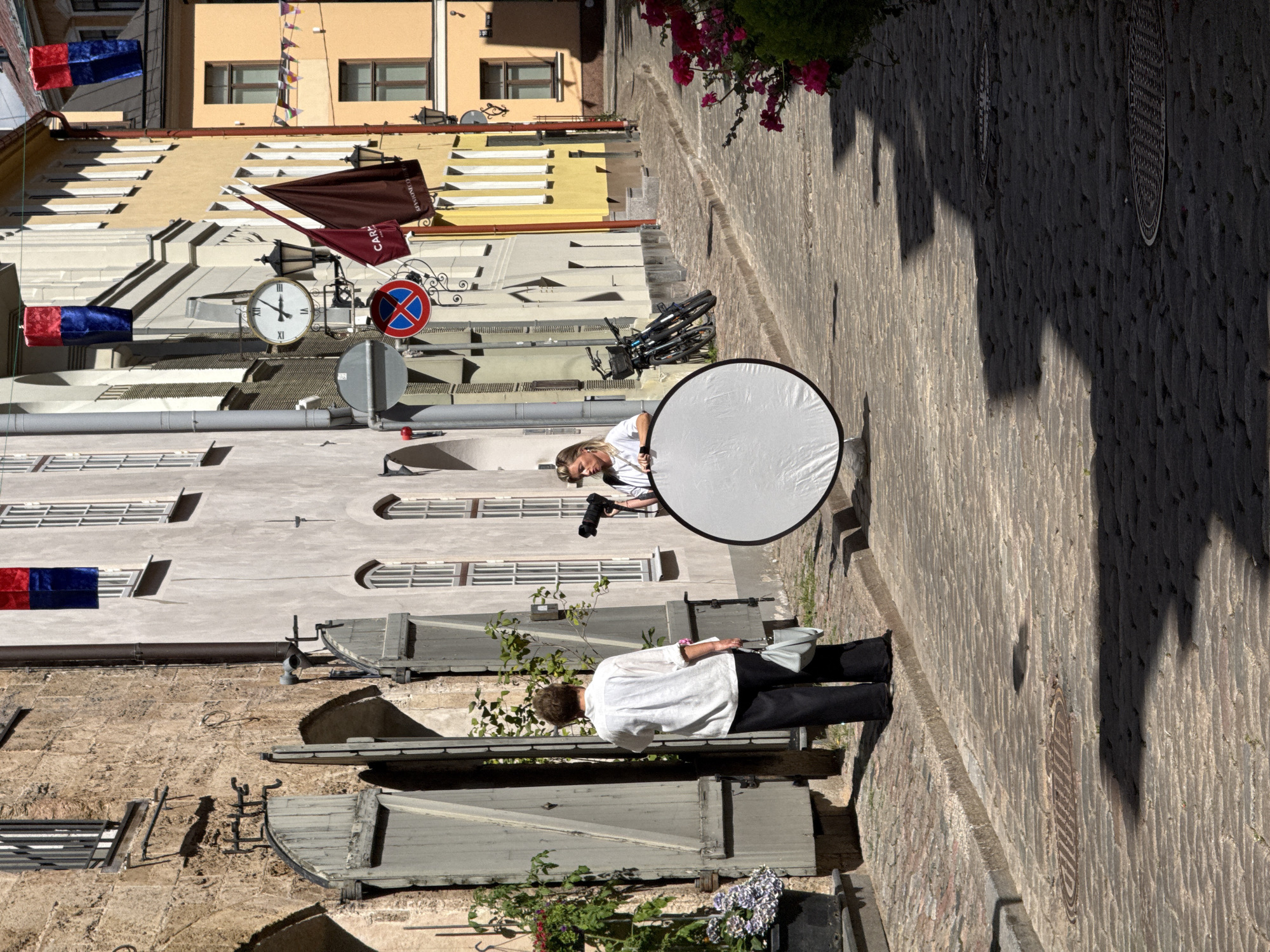The image depicts a narrow, cobblestone street in Riga, Latvia, possibly in the Old Town. The architecture suggests historical buildings with textured stone facades and arched doorways. Sunlight casts sharp shadows on the street, indicating a bright, sunny day.
In the foreground, a woman dressed in a white shirt and black pants stands facing away from the camera, holding a light blue handbag. She is positioned in front of an open, weathered wooden shuttered entrance.
Further down the street, another woman, wearing a white top with a striped neckline, is holding a camera and a large, circular reflector. She appears to be engaged in a photoshoot, with the reflector positioned to bounce light.
Several bicycles are parked against a building in the background. Decorative flags or banners hang from some of the buildings, adding a touch of color to the scene. A clock tower is visible on a building to the right of the street. The overall impression is a candid moment captured during an outdoor photography session on a picturesque, historic street.