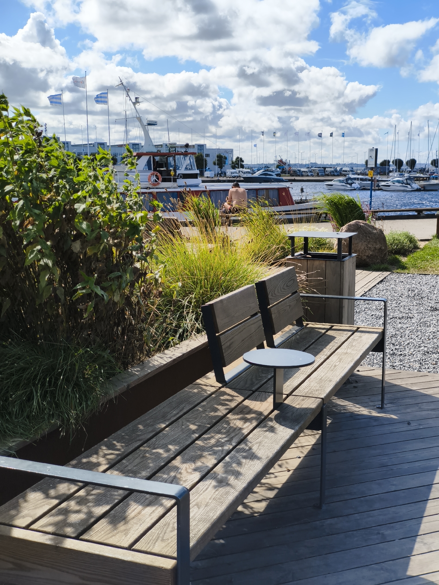 The image shows a waterfront scene in Tallinn, Estonia. In the foreground, a wooden bench with metal armrests faces a body of water. A small, round side table is attached to the bench. To the left of the bench, lush green foliage grows in a raised planter. Behind the bench, a wooden trash receptacle with a metal top stands next to a large, rough-hewn rock. In the background, a harbor filled with various boats and yachts is visible. A larger boat with a red and white hull and flags on its mast is docked to the left. Further in the distance, buildings and more boats line the harbor. The sky is a vibrant blue with scattered, fluffy white clouds. The scene appears to be a public area, possibly a park or promenade, adjacent to the harbor. A person, unclothed from the waist up, is seated on a structure in the middle ground, facing away from the camera towards the water.