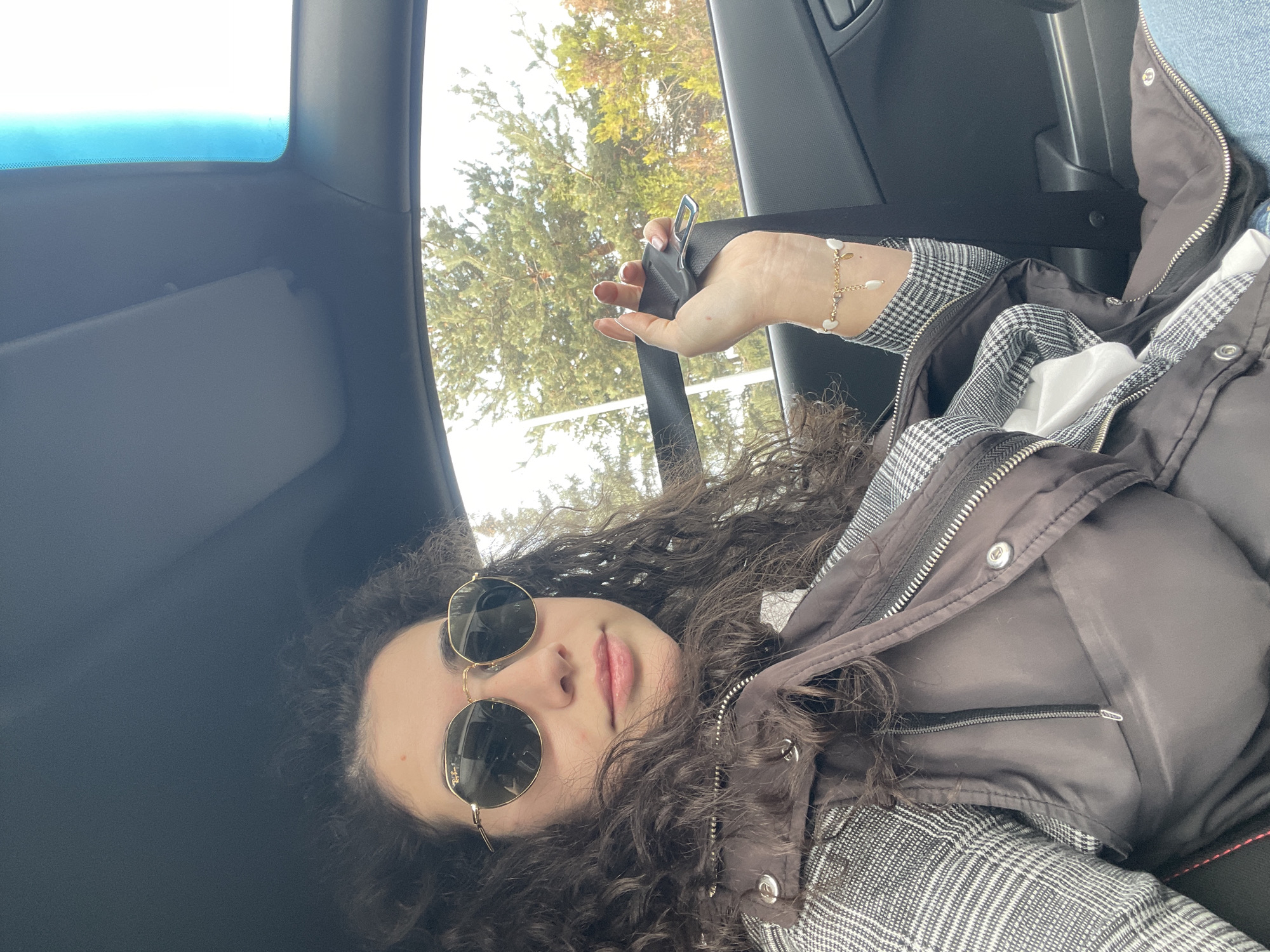 A young woman with long, dark curly hair lies reclined in what appears to be the front passenger seat of a car, looking towards the camera. She wears round, gold-rimmed sunglasses with "Ray-Ban" visible on the lens, obscuring her eyes. Her lips are slightly parted, conveying a relaxed or pensive expression. She is dressed in a plaid patterned shirt, visible beneath a dark, likely brown or black, puffer vest with metallic snap buttons and zippers. A delicate gold bracelet adorned with white, bead-like charms is visible on her left wrist, and her hand rests near the car's seatbelt buckle. The car's interior, including a grey roof liner, sun visor, and an A-pillar, is seen above and around her. Through the side window, bright natural light illuminates green and brown trees, indicating a daytime outdoor setting in Sofia, Bulgaria, likely with clear weather.