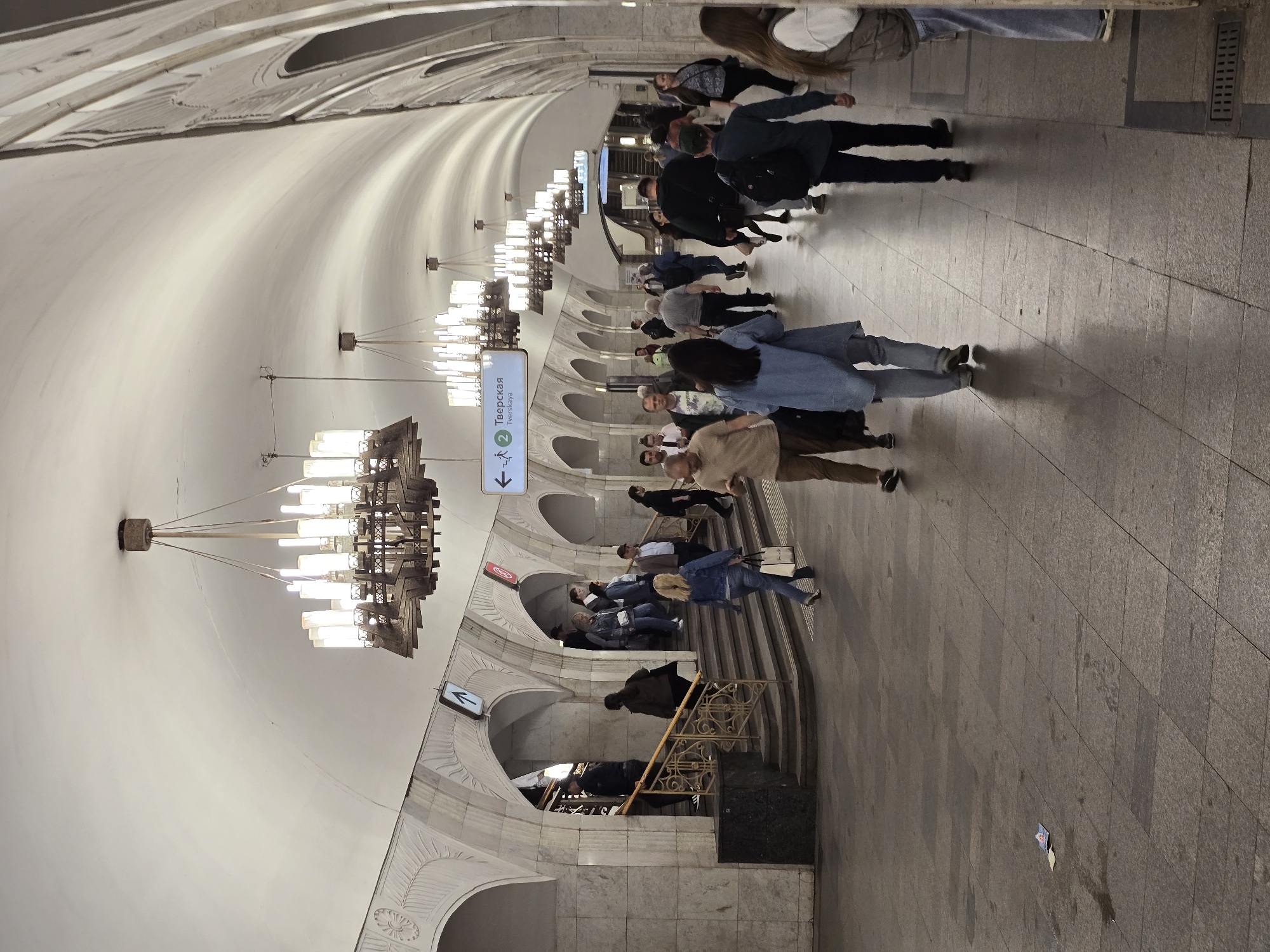 This image shows an interior view of the Tverskaya station of the Moscow Metro. The station's architecture features arched entrances, ornate lighting fixtures, and marble walls. People are walking and standing in the station, and a sign indicates the station name in Russian, "Тверская" (Tverskaya).