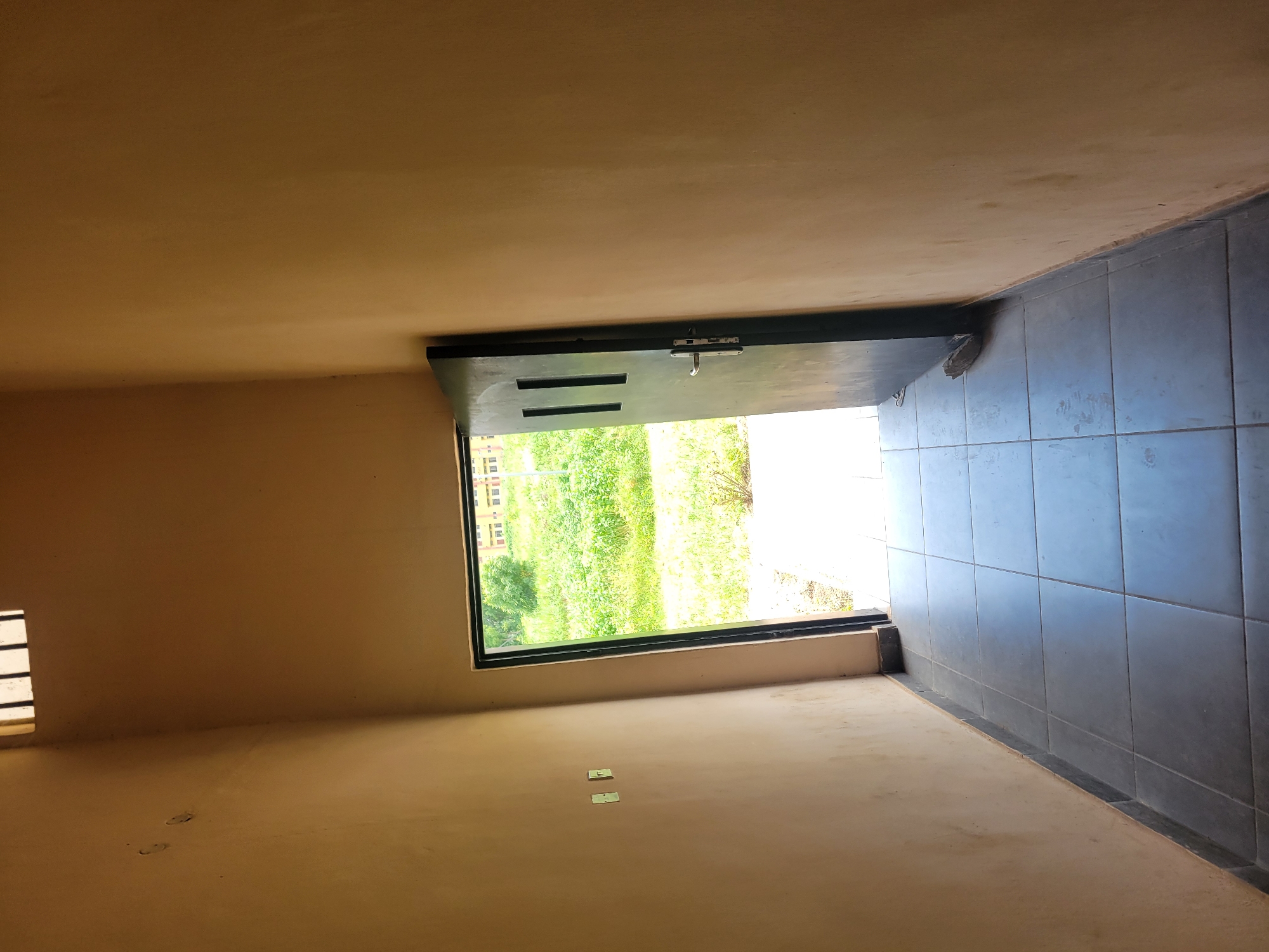 An interior shot shows a narrow hallway with tiled floor leading to an open dark gray door. The walls are painted a light brown color and there are two white electrical outlets on the left wall. The door frame is dark gray and the door itself has two rectangular windows with black frames. Beyond the open door, there is a bright outdoor scene with green grass and a building in the background. The hallway has a tiled floor made of large, square, gray tiles. The top of the frame shows a slatted window.