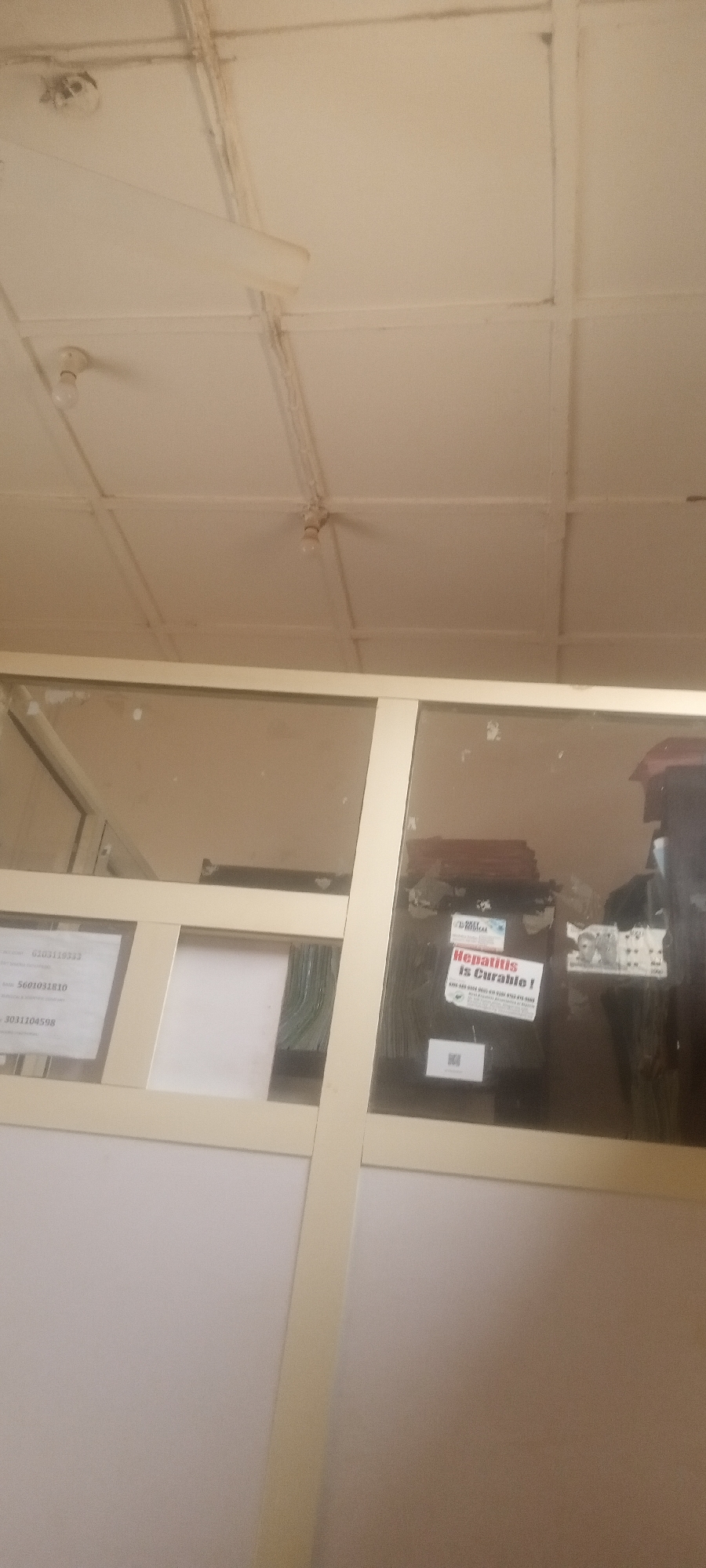 The image shows an interior view, possibly within a building, featuring a partition with glass panels. The upper portion of the frame appears to be reflective, revealing objects behind it, which might be office equipment and papers. The lower portion of the partition appears to be opaque, with a light beige or off-white color. On one of the glass panels, there's a sign that reads "Hepatitis is Curable!". Also visible are phone numbers written on a sheet of paper.
Above the partition is a white, textured ceiling with visible light fixtures and possibly a ceiling fan. The scene seems to be indoors, possibly in an office setting.
Given the prompt's information, this likely takes place in a building located in Yelwa, Nigeria, where the partition functions as a customer service interface.