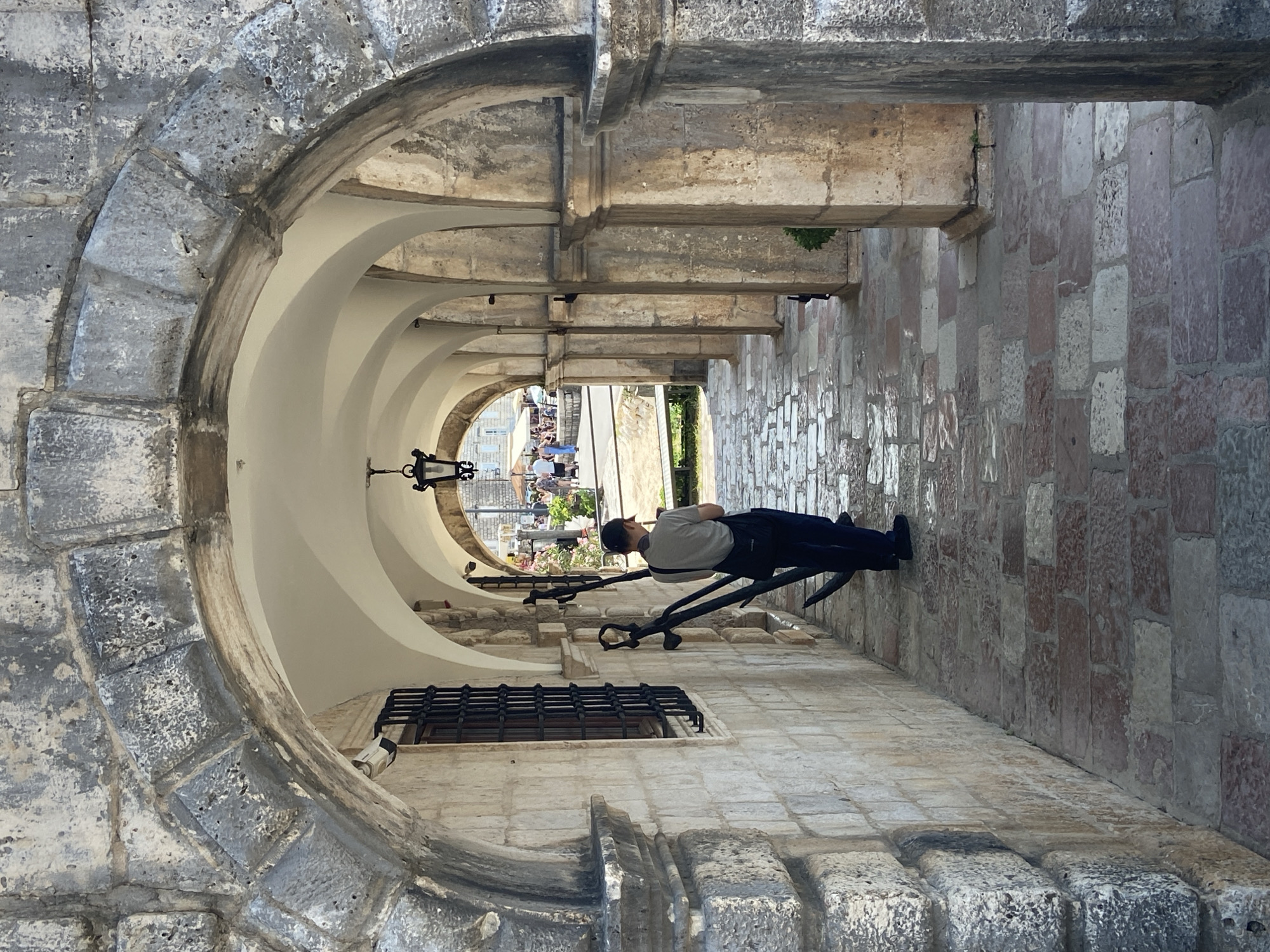 A person is standing under a stone arcade in Perast, Montenegro. The arcade features a series of arches supported by stone columns, with a tiled floor. The person, seen from behind, is wearing a light-colored shirt, dark trousers, and a dark shoulder bag. Two large anchors are leaning against the wall to the left of the person. Beyond the arcade, an outdoor area with tables and chairs, and other people, is visible. The architecture suggests a historical setting, likely a waterfront area.