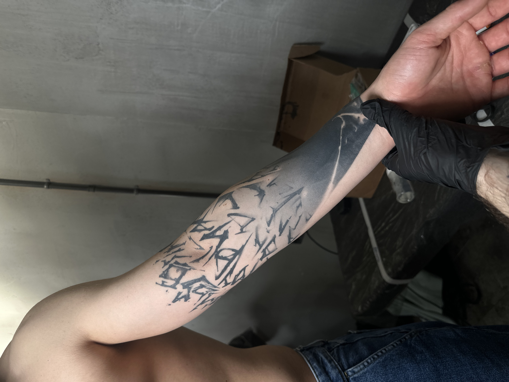 A person lies on their back, wearing blue jeans, with their bare torso partially visible. Their left arm is extended and tattooed with a black design consisting of abstract geometric shapes and a solid black section towards the wrist. A hand wearing a black latex glove holds the tattooed arm near the wrist. The scene appears to be indoors, under artificial lighting, with a gray concrete wall in the background. A black tube, a cardboard box, and a partially obscured object, possibly part of a table or workstation, are visible in the background. The location is not identifiable as Moscow, Russia, without additional information.