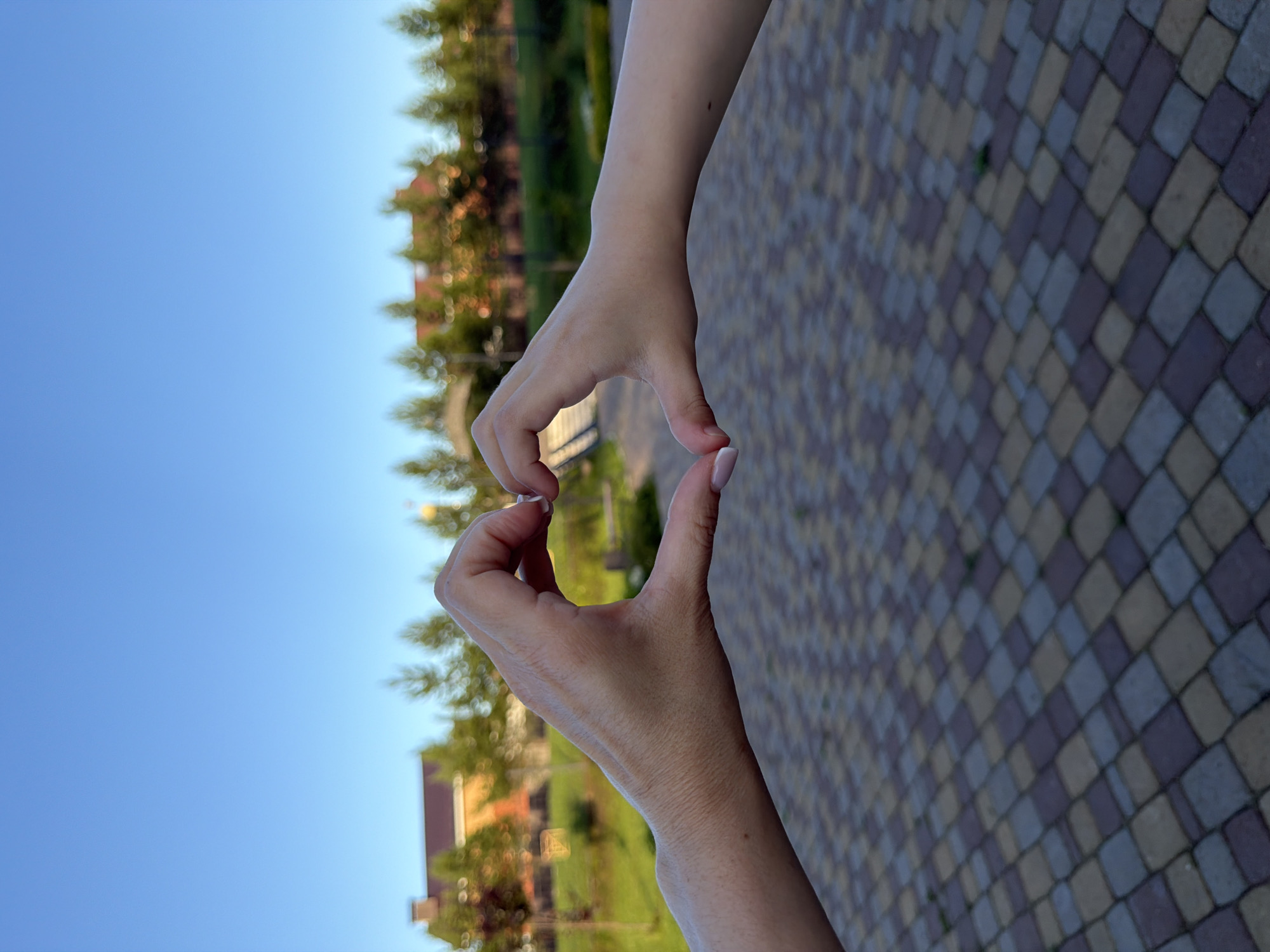 The image shows a close-up of two hands forming a heart shape against a backdrop of a clear blue sky, green trees, and a patterned paved surface. The hands appear to belong to a person or people, and they are the central focus of the image. The heart shape is created by bringing the thumbs and index fingers of both hands together. The lighting suggests it is daytime, likely sunny given the bright sky. The background is slightly blurred, indicating a shallow depth of field, which further emphasizes the hands. The paved surface is made of interlocking rectangular stones in varying shades of grey, brown, and purple. The trees in the background are lush and green, and some appear to be coniferous. Hints of a building with a reddish roof can be seen among the trees. The overall mood conveyed by the image is positive and affectionate, suggested by the universal symbol of a heart. No text is visible in the image. The location context provided is Шмидтово, Russia.