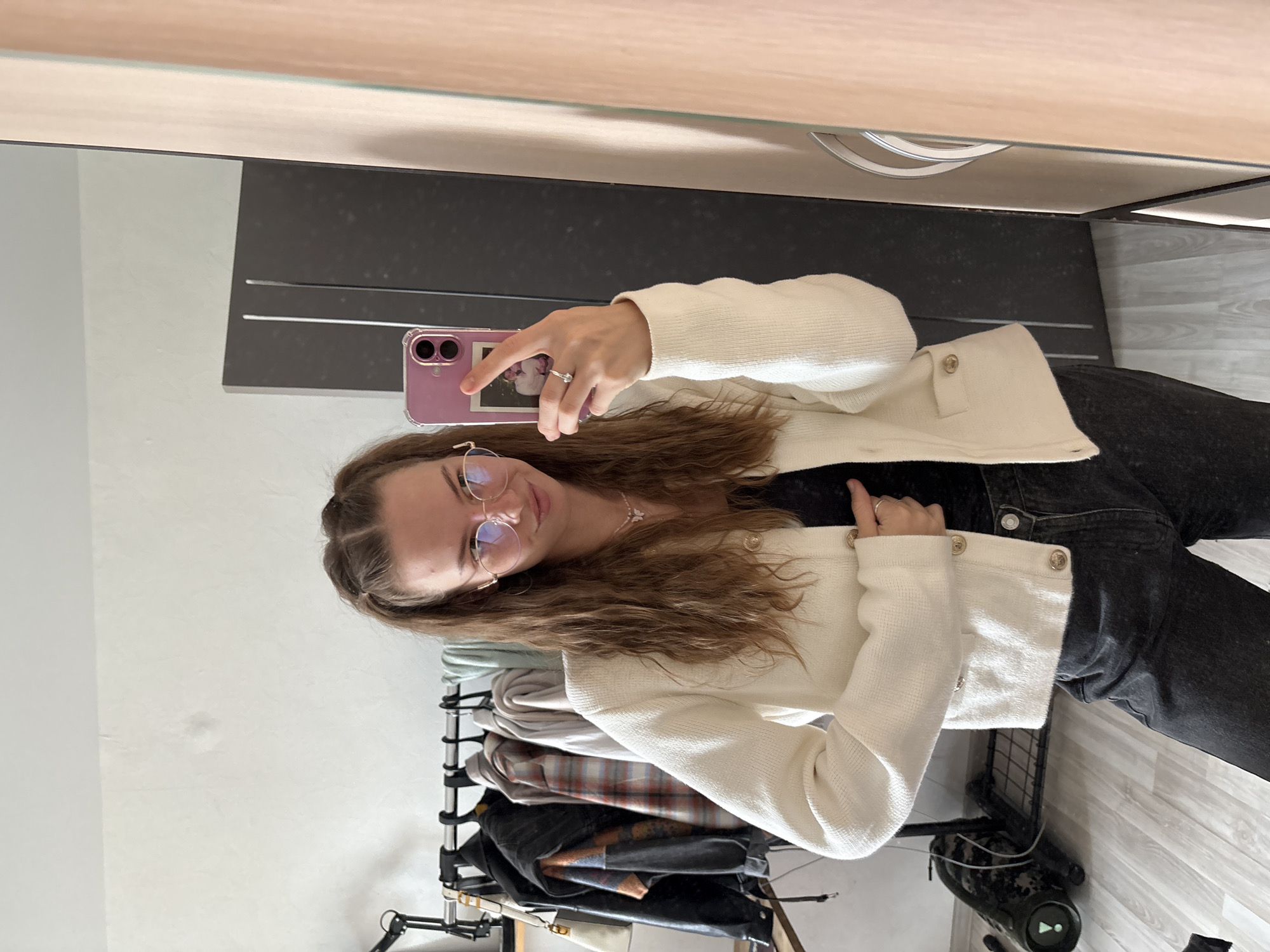 A young woman with long, wavy brown hair tied in pigtails is taking a mirror selfie in a room. She is wearing a white, textured cardigan over a black top and dark wash jeans. She has on round, gold-rimmed glasses and a delicate necklace with a butterfly pendant. Her left hand is resting on her stomach, and her right hand is holding a pink phone, on which a small photo is visible. She is wearing a ring on her right ring finger. In the background, there is a clothing rack with several items of clothing, including a black leather jacket and plaid shirts. To the right, a tall, dark wardrobe is partially visible, and a mirror reflects the room. The floor is light-colored wood. The scene appears to be a bedroom or dressing area, likely in a residential apartment, not a public space in the city of Perm, Russia.