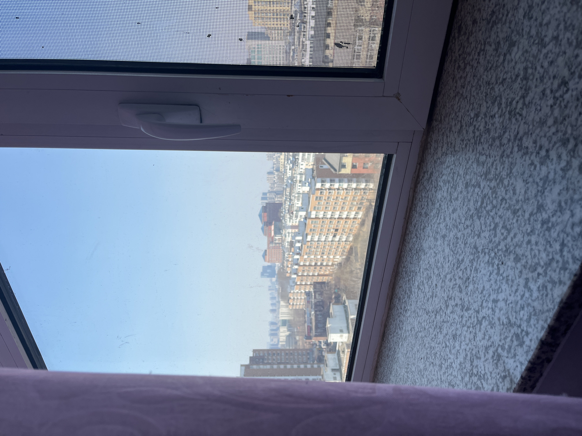The image depicts an elevated outdoor view of an urban residential area in Changchun City, China, observed through an interior window on a clear day. The foreground shows a speckled grey and white window sill extending across the bottom, with a light purple curtain partially obstructing the left side of the frame. A white uPVC window frame with a white handle on the right vertical mullion is prominent. The window's glass pane, visible on the left, is smudged, while the right section features a dark mesh screen with visible dirt and debris.
Beyond the window, a dense urban landscape unfolds under a bright, cloudless blue sky. The midground features numerous multi-story residential buildings, primarily displaying light brown or orange-brick facades with white trim. Bare deciduous trees are interspersed between these structures, suggesting a late autumn or winter season. In the distant background, taller, darker high-rise buildings contribute to the city skyline. No individuals are present in the scene, and no specific actions are depicted.
