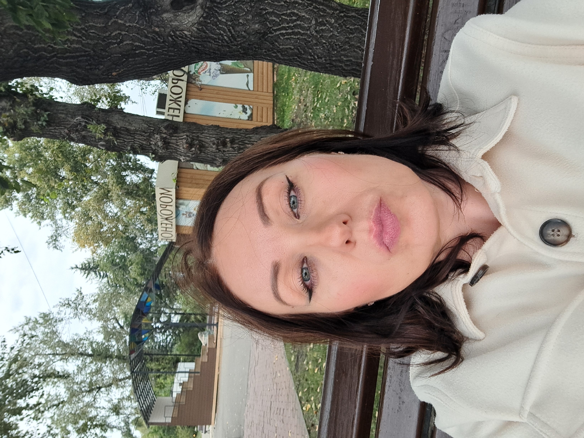 A woman is sitting on a park bench, looking at the camera with her lips pursed. She has dark hair and is wearing a cream-colored jacket with two buttons. In the background, there is a small stage with tiered seating and a sign that reads "МОРОЖЕНОЕ" (ice cream) on a building. Trees and grass are visible in the park. The location is the Kirov Park in Irkutsk, Russia.
