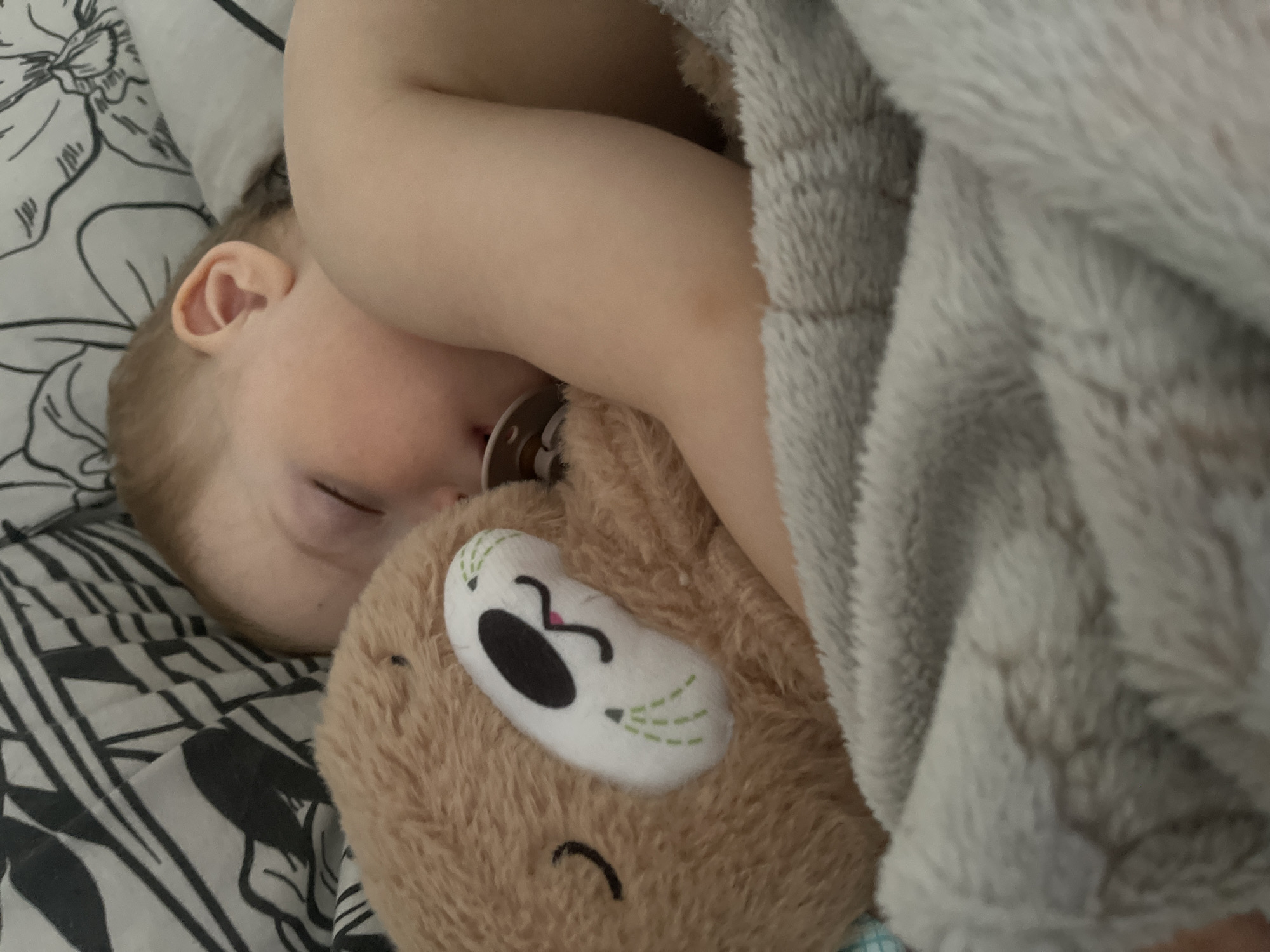 A close-up shot depicts a sleeping child with light-colored skin and hair. The child is clutching a stuffed, brown, plush toy with a white eye patch and stitched detailing. A pacifier is in the child's mouth. The child is lying on a black and white patterned pillowcase and is partially covered by a gray fuzzy blanket. The scene takes place indoors. The location of Izhevsk, Russia, is not identifiable from the given image.