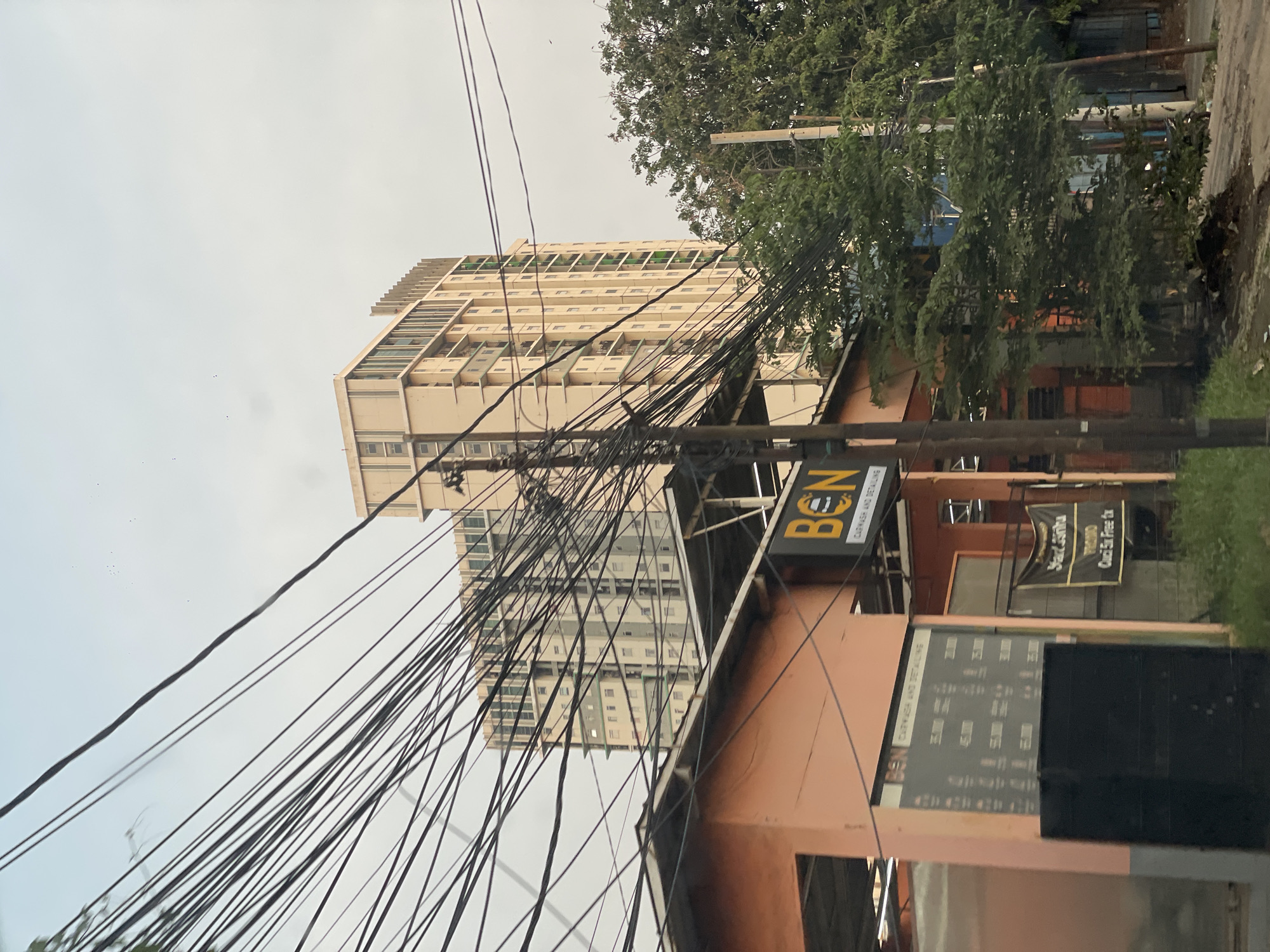 The image captures a street scene in North Jakarta, Indonesia, dominated by a tangle of overhead power lines. The focus is on a building, possibly a multi-story apartment complex, visible behind the power lines. In the foreground, there is a car wash and detailing business called "BEN" with a sign displaying prices and services. The sky is overcast. The scene appears to be taken from a vehicle, perhaps while driving. There are no visible people.