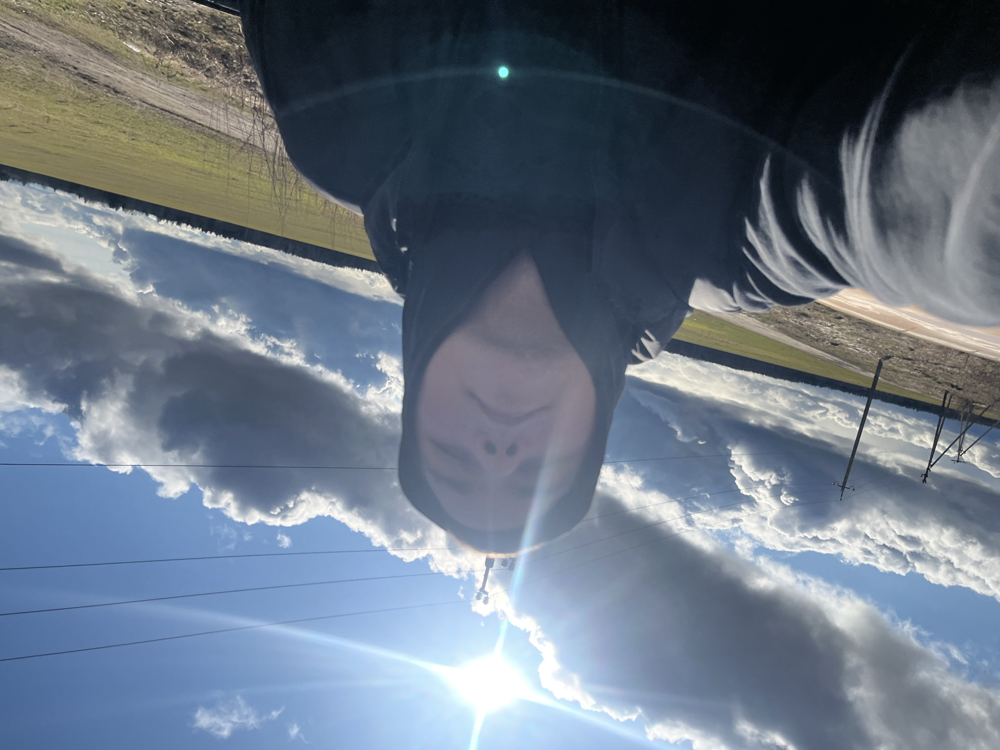 The image shows a person wearing a dark hooded jacket. The background features a blue sky with white and grey clouds, power lines, and utility poles. In the distance, a field or grassy area is visible. The photo appears to be taken from a low angle, looking upwards toward the sky and person. The city of Ямуга, Russia, is given as the location, but this is not verifiable from the image itself.