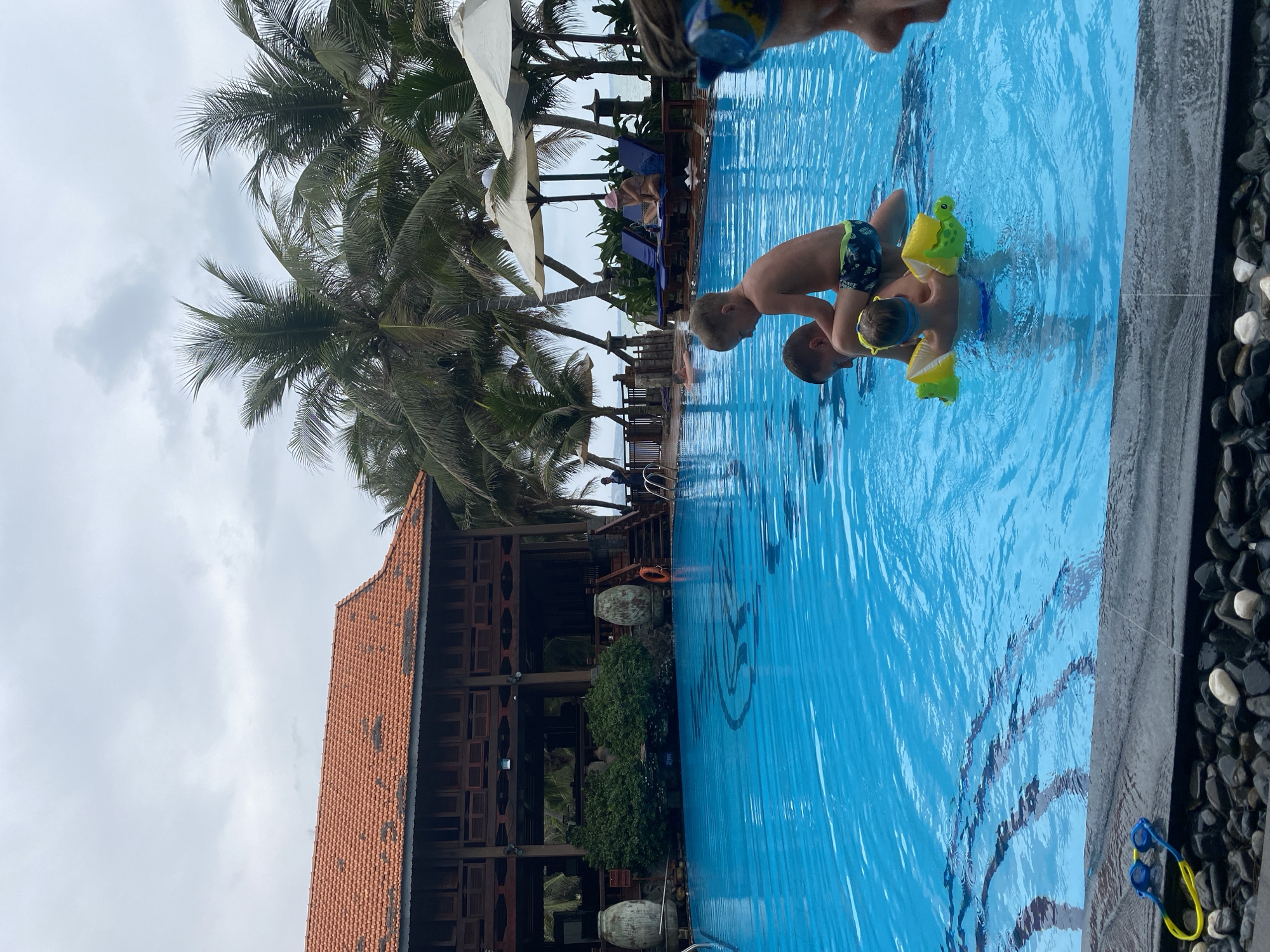 This image, captured in Phu Thuy Ward, Vietnam, depicts an outdoor swimming pool area under an overcast sky. In the bright blue pool, two young boys are playing, seemingly supported by an adult whose head and shoulders are partially visible at the top of the frame. One boy wears green and black swim trunks, yellow arm floaties, yellow goggles, and holds a yellow toy turtle. The other boy, also in swim trunks, is close by.
The pool has visible steps leading into the water, and its edge is lined with dark and white pebbles. A pair of blue and yellow goggles rests on these pebbles. The bottom of the pool features a faint blue, stylized pattern and the text "2m," indicating the water depth.
The background showcases numerous tall palm trees with lush green fronds. To the left, a multi-story building with a distinctive terracotta-tiled roof and dark wooden structure, possibly featuring shutters or balconies, establishes a resort-like setting. Beyond the pool and trees, lounge chairs and umbrellas are visible, and a distant body of water, likely the ocean or sea, can be seen, reinforcing the coastal location. The scene conveys a relaxed, recreational atmosphere.