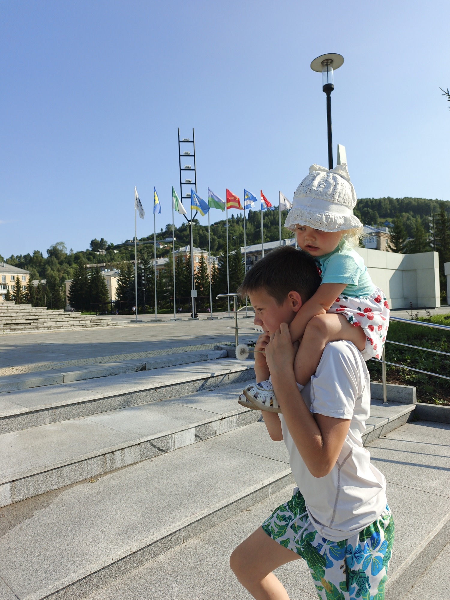 In Zlatoust, Russia, a young boy carries a toddler on his shoulders up a wide outdoor staircase. The boy wears a white t-shirt and green and white floral shorts, while the toddler is dressed in a light blue shirt and a white bonnet. The background reveals a clear blue sky, a flagpole with multiple flags, and buildings nestled amongst lush green trees on a hillside. The setting appears to be a public space within the city.