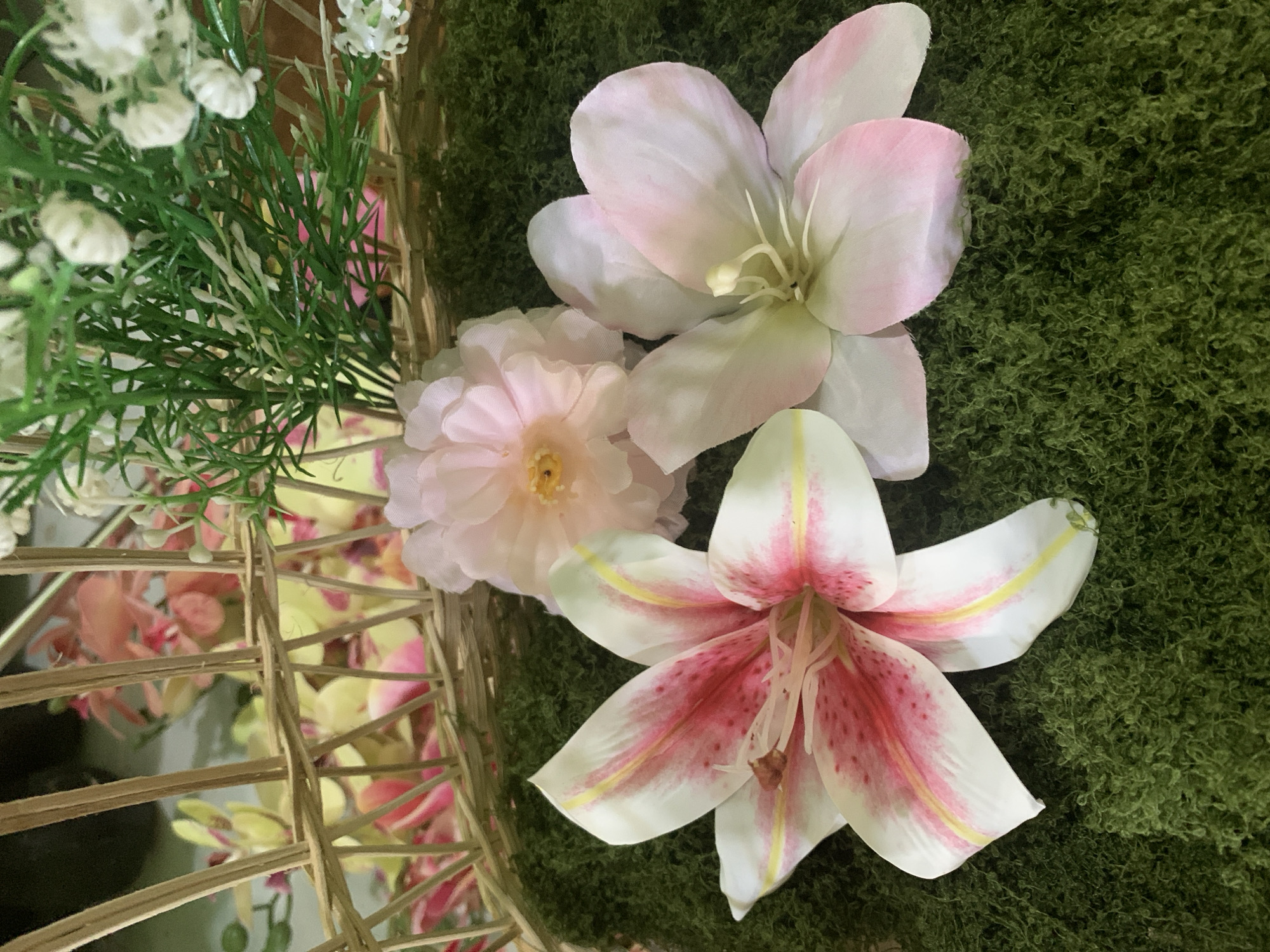 A still life image captures a decorative arrangement of artificial flowers on a bed of dark green, textured moss. In the foreground, two lilies are prominent. The lily on the left is white with vibrant pink speckles and streaks, and yellow accents along its petals. The lily on the right is predominantly pale pink with white undertones. Behind these, a lighter pink, ruffled flower is partially visible. The arrangement is set within a wicker basket, the weave of which is visible in the upper portion of the frame. This basket contains more artificial flowers in shades of pink and yellow, and a sprig of green foliage with small white buds. The background is blurred and dark. The scene is indoors, with soft lighting. There is no indication of a specific location within Kuala Pembuang, Indonesia.