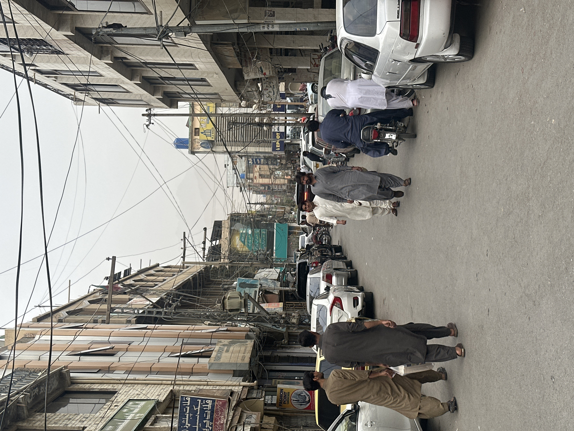 The image shows a street scene in Quetta City Tehsil, Pakistan. The street is lined with multi-story buildings on both sides, displaying various storefronts and signage. A number of people are walking and standing on the street. Several cars are parked and driving on the road, with a motorcycle also visible. Overhead, a complex network of electrical wires crisscrosses the sky. The overall atmosphere suggests a busy, commercial area within a city.