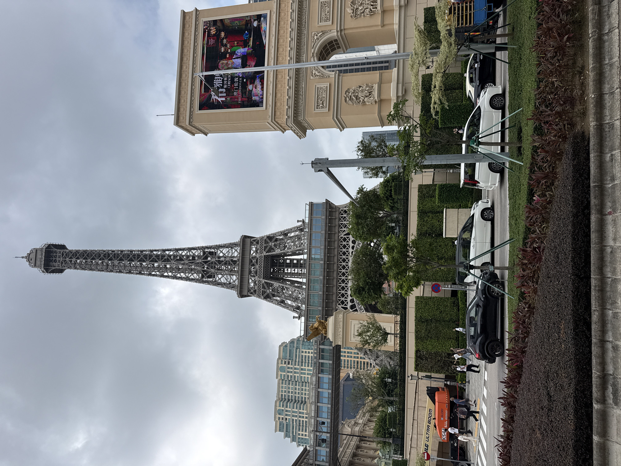 The image shows a partial view of the Parisian Macao integrated resort in Macao, China. The Eiffel Tower replica is the main subject, extending from the lower left upward. The Parisian Arc de Triomphe facade is partially visible on the upper right with a billboard atop. Several cars are parked along the roadside to the right of the image. People are seen crossing a crosswalk on the lower right, near a construction lift. Buildings and greenery are also visible, and the sky is overcast.