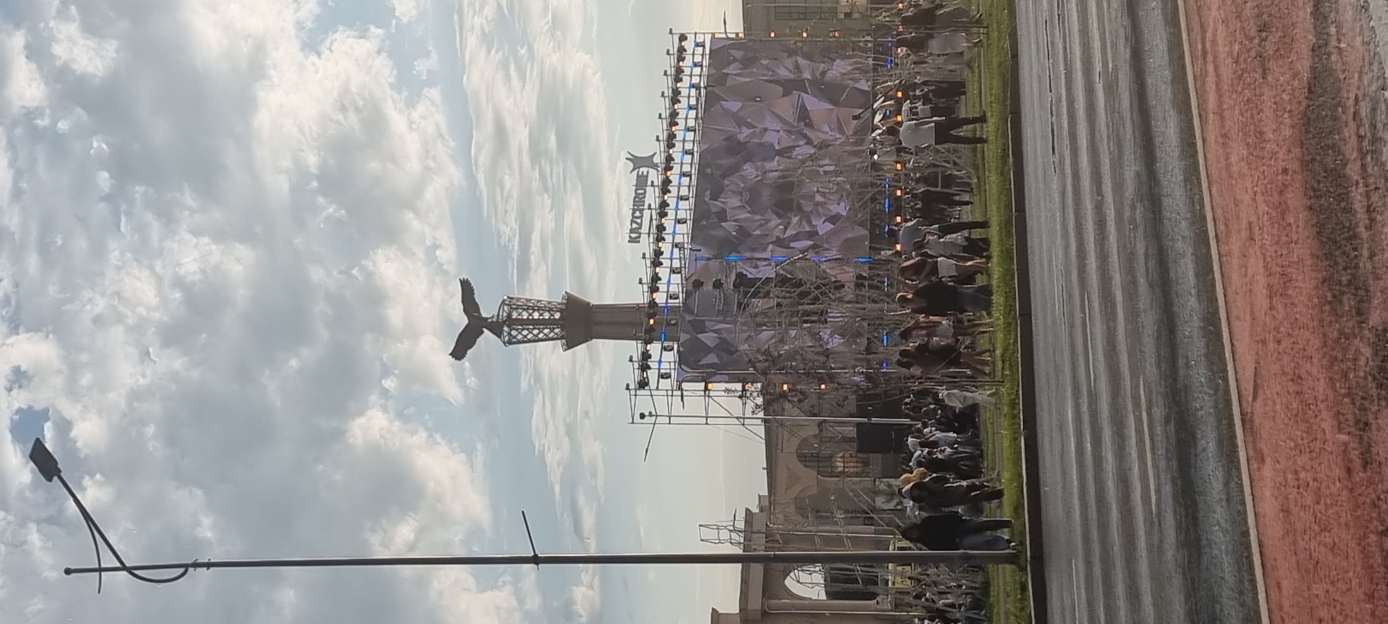 In Aktobe, Kazakhstan, a wide street with wet asphalt and faint white lane markings borders a grassy area where a crowd of people are gathered. In the background, a large outdoor stage is set up with a massive LED screen displaying a geometric pattern. Above the stage, a prominent statue of an eagle atop a decorative tower stands against a cloudy sky. A tall street lamp with a curved arm is visible to the left of the frame. The overall scene suggests an outdoor event or festival is taking place.