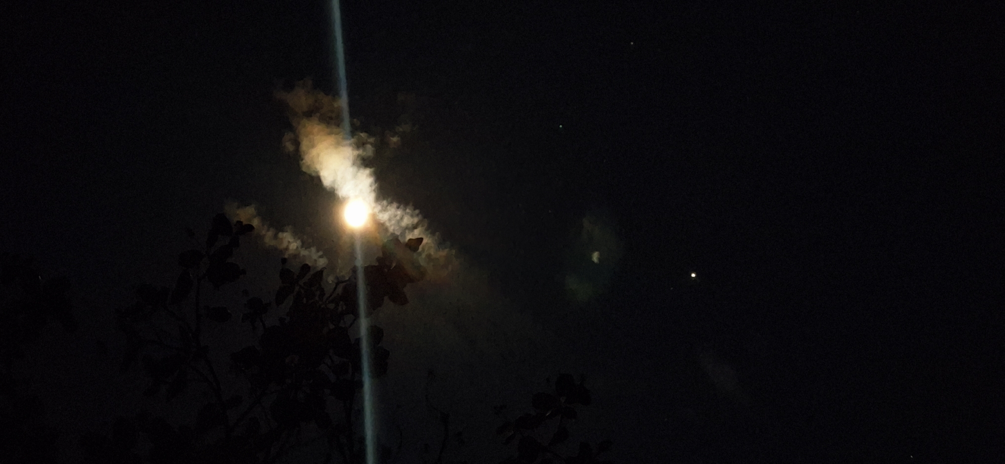 A nighttime photograph captured in Vallecito, Venezuela shows the moon shining brightly through a layer of clouds, with a distinct light streak emanating from it. In the foreground, the silhouette of tree branches and leaves is visible, suggesting a natural setting. In the distance, a faint green crescent moon, possibly representing the same object, is seen to the right of the brighter, obscured moon. Two other points of light, possibly stars, are scattered across the night sky.