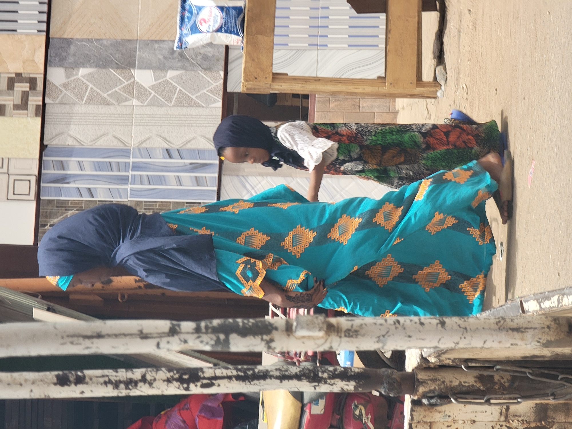 In an outdoor scene in Maiburiji, Nigeria, an adult wearing a dark blue hijab and a long, teal dress with orange geometric patterns stands next to a child. The child, also wearing a dark head covering and a white top, stands closer to a display of stacked tiles. The backdrop includes various patterned tiles, a wooden frame holding additional tiles, and a glimpse of an unpaved surface. In the foreground, a section of a weathered, metal structure partially obscures the lower part of the view. The adult's hand is visible, adorned with henna designs.