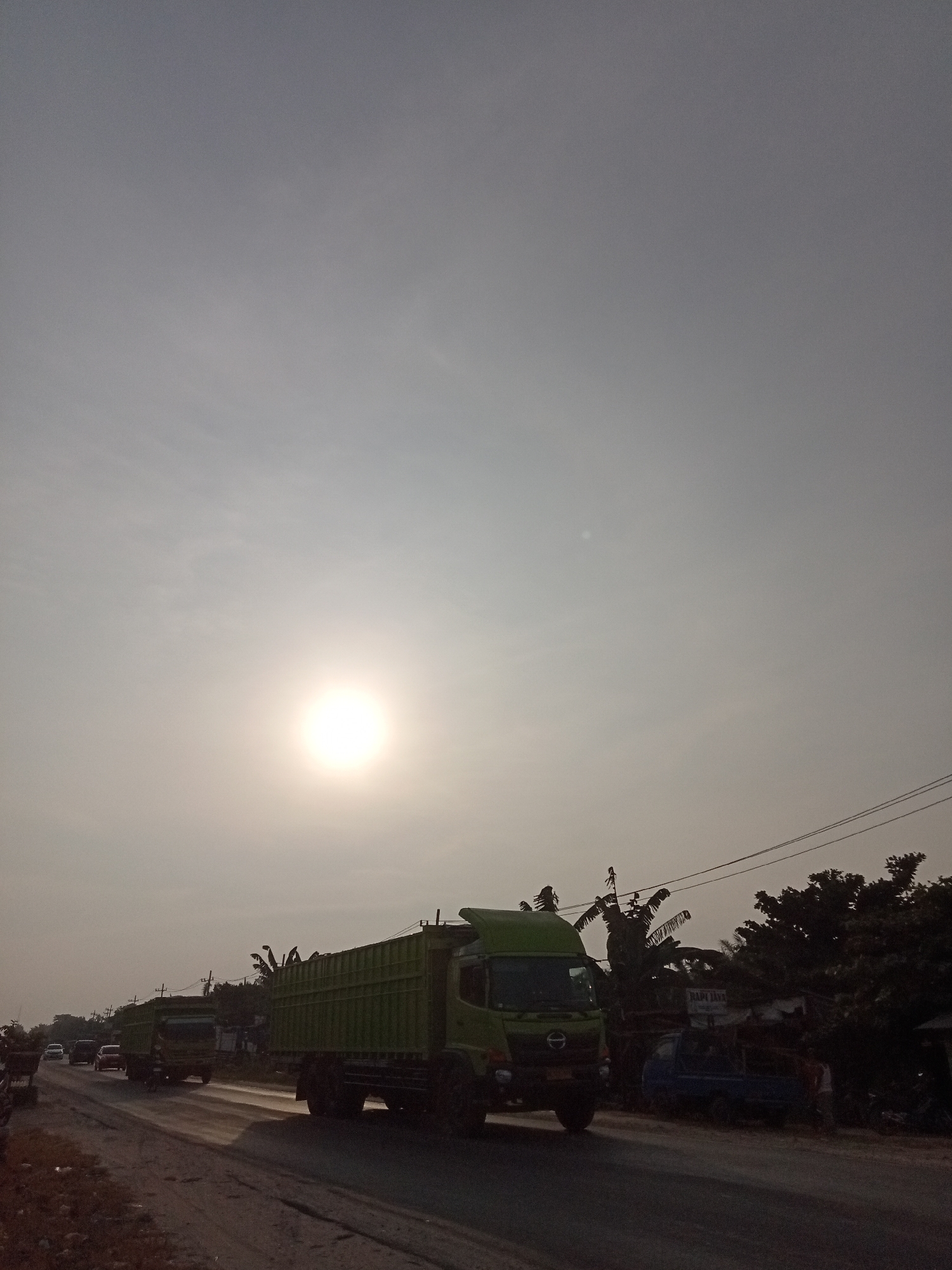 The image depicts a street scene in Kubang Jaya, Indonesia, during what appears to be late afternoon or early morning, with the sun low in the hazy sky. The foreground is dominated by a dirt or gravel road with tire tracks. On the road, a large green truck is moving away from the viewer towards the right. To the left, several other vehicles, including cars and possibly another truck, are also traveling on the road. In the background, there are trees and some structures, including what looks like a building with a sign that is partially legible, possibly reading "NAPI JAYA". Power lines are visible overhead. The overall atmosphere is somewhat muted due to the haze and the low sun angle. There are no clearly identifiable individuals captured in detail.