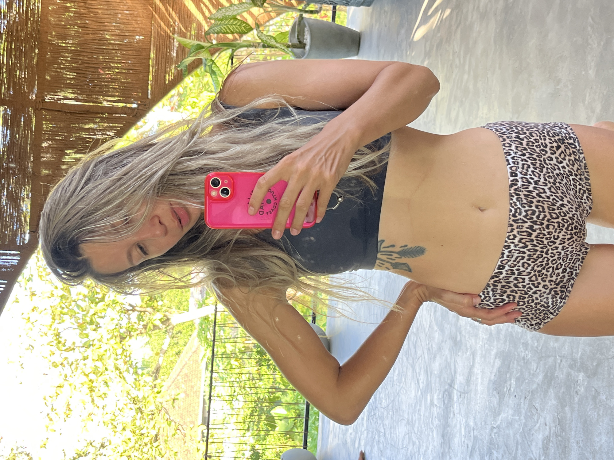 A woman is taking a selfie in an outdoor setting, possibly a balcony or terrace, in Weligama, Sri Lanka. She has blonde hair and is wearing a dark sports bra and leopard-print shorts. She is holding a pink smartphone in her right hand, which is positioned to capture the photo. Her left hand is on her hip. In the background, lush green foliage and trees are visible, suggesting a tropical environment. The architecture appears to be made of natural materials, such as bamboo, and there are potted plants. The lighting indicates it is daytime.