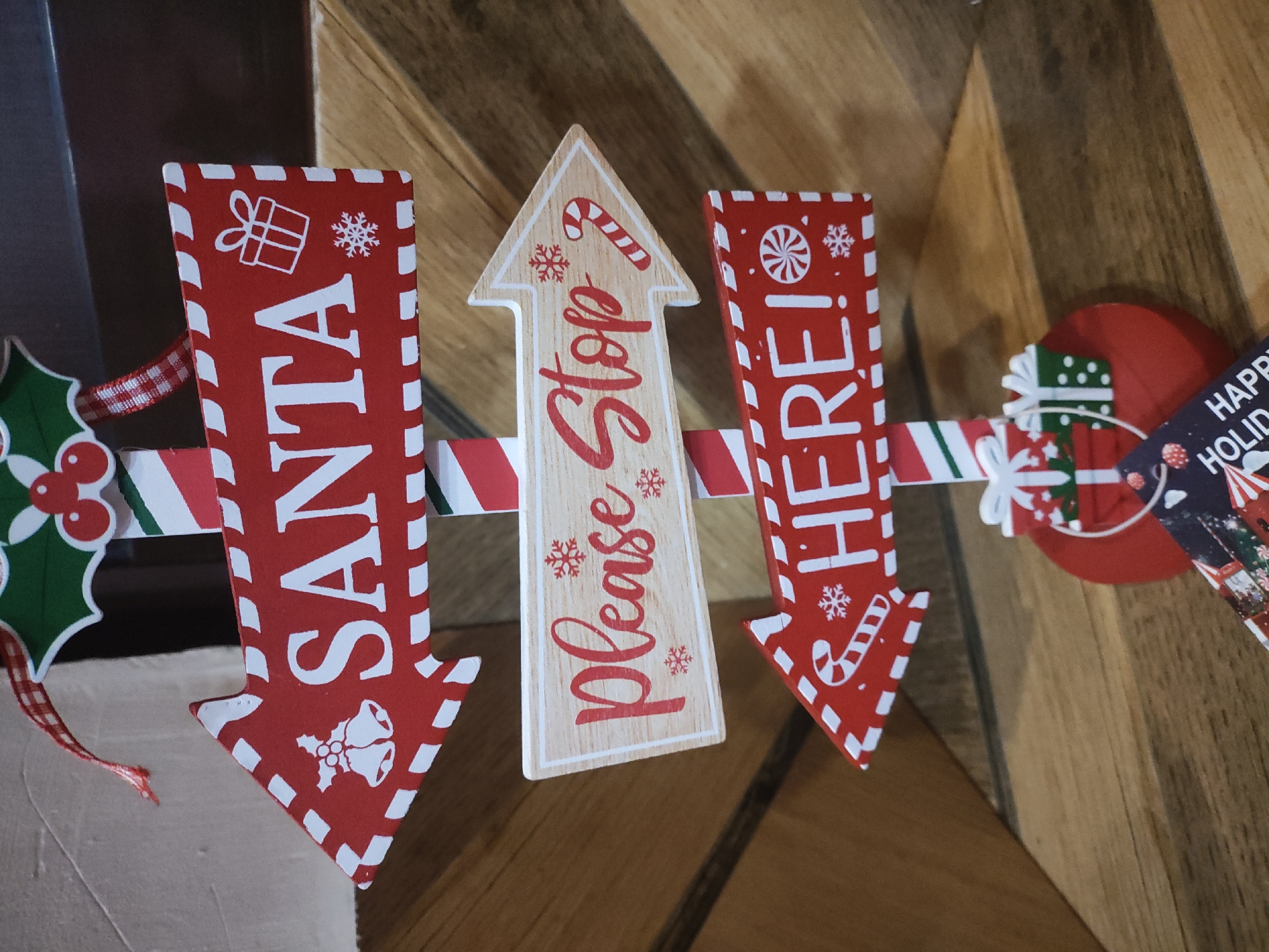 Three Christmas-themed arrow signs are connected by a candy cane-striped ribbon. The sign on the left is red and white and says "Santa", the middle sign is tan and says "Please Stop", and the sign on the right is red and white and says "Here!". There are small Christmas ornaments dangling from the ribbon. The background is a wooden surface.