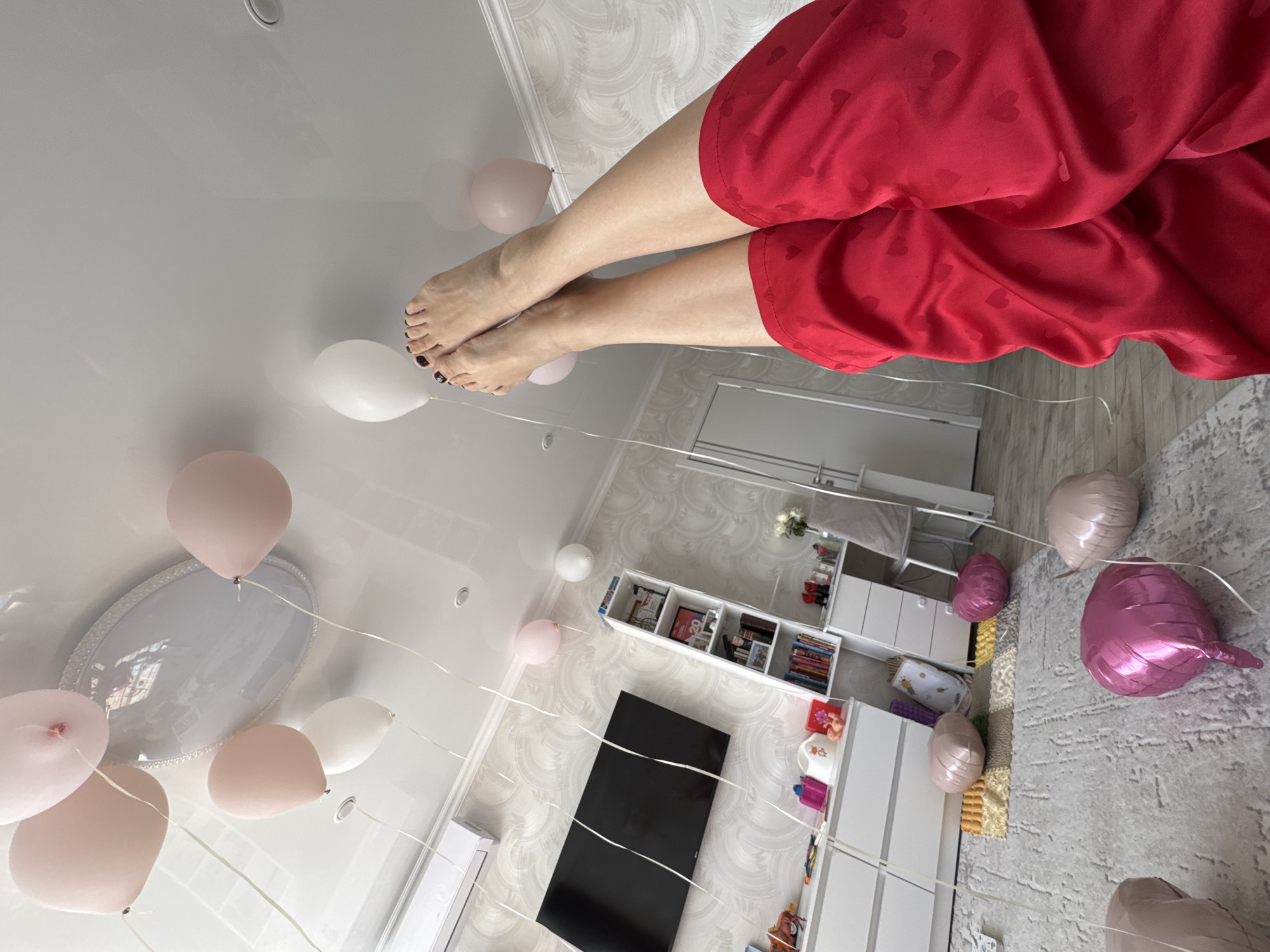 The image shows a room decorated with balloons in Sochi, Russia. The view is from an elevated angle, as if looking down on the scene. A person wearing red pajama pants with a heart pattern is visible, with their legs extended towards the ceiling. Pink and white balloons are scattered throughout the room, tied with white strings. The room contains a bathtub with a decorative tray, a television, shelving, and other furniture. The walls have a patterned wallpaper. The floor is carpeted in the area away from the bathtub.