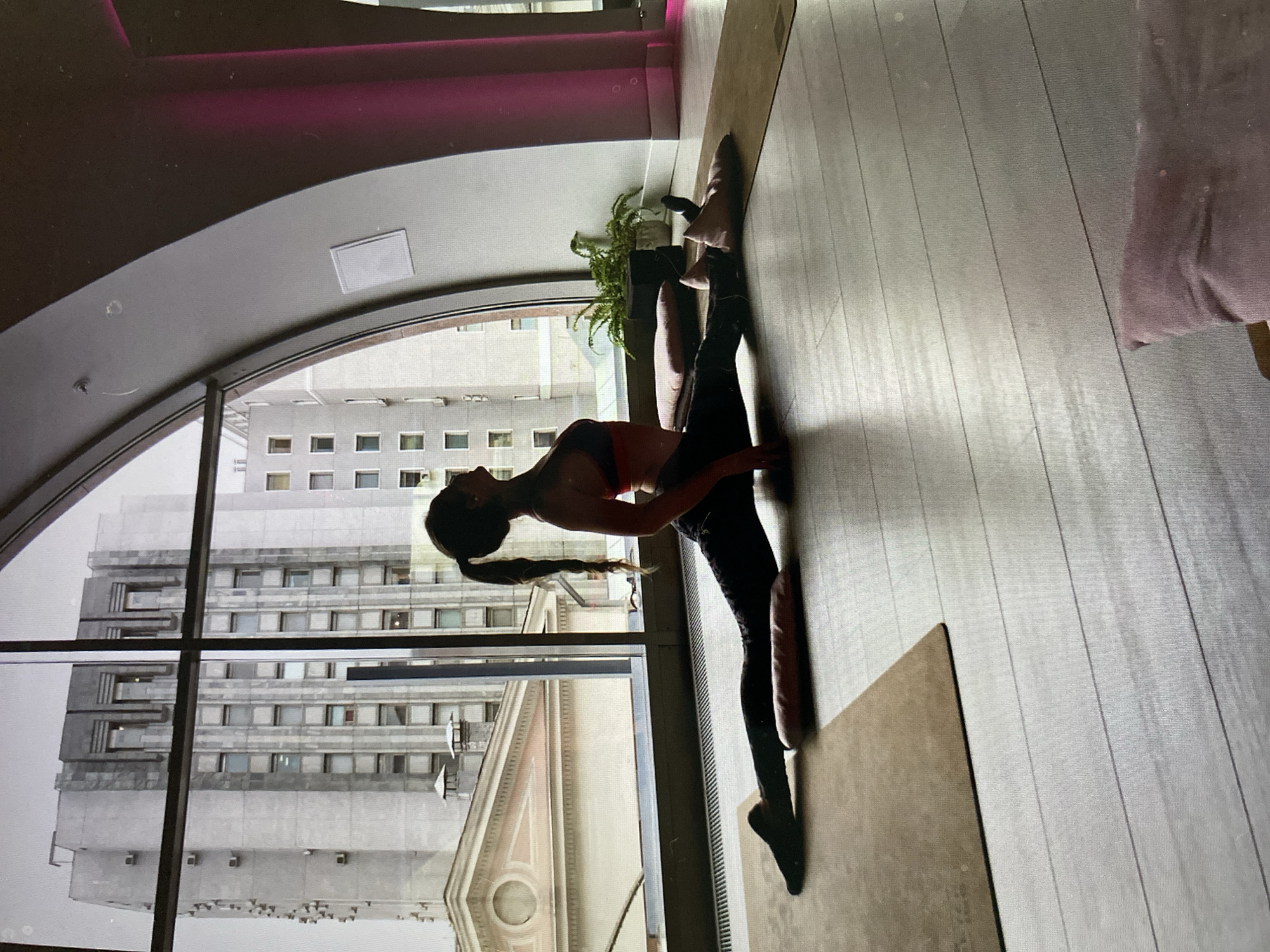 A woman is practicing a yoga split on a mat in a yoga studio. She is wearing black leggings and a sports bra. The studio has large windows that overlook a city with tall buildings. A plant is visible in the background. The lighting in the studio is dim, with pink accents in the background. The floor is made of light-colored wood planks.