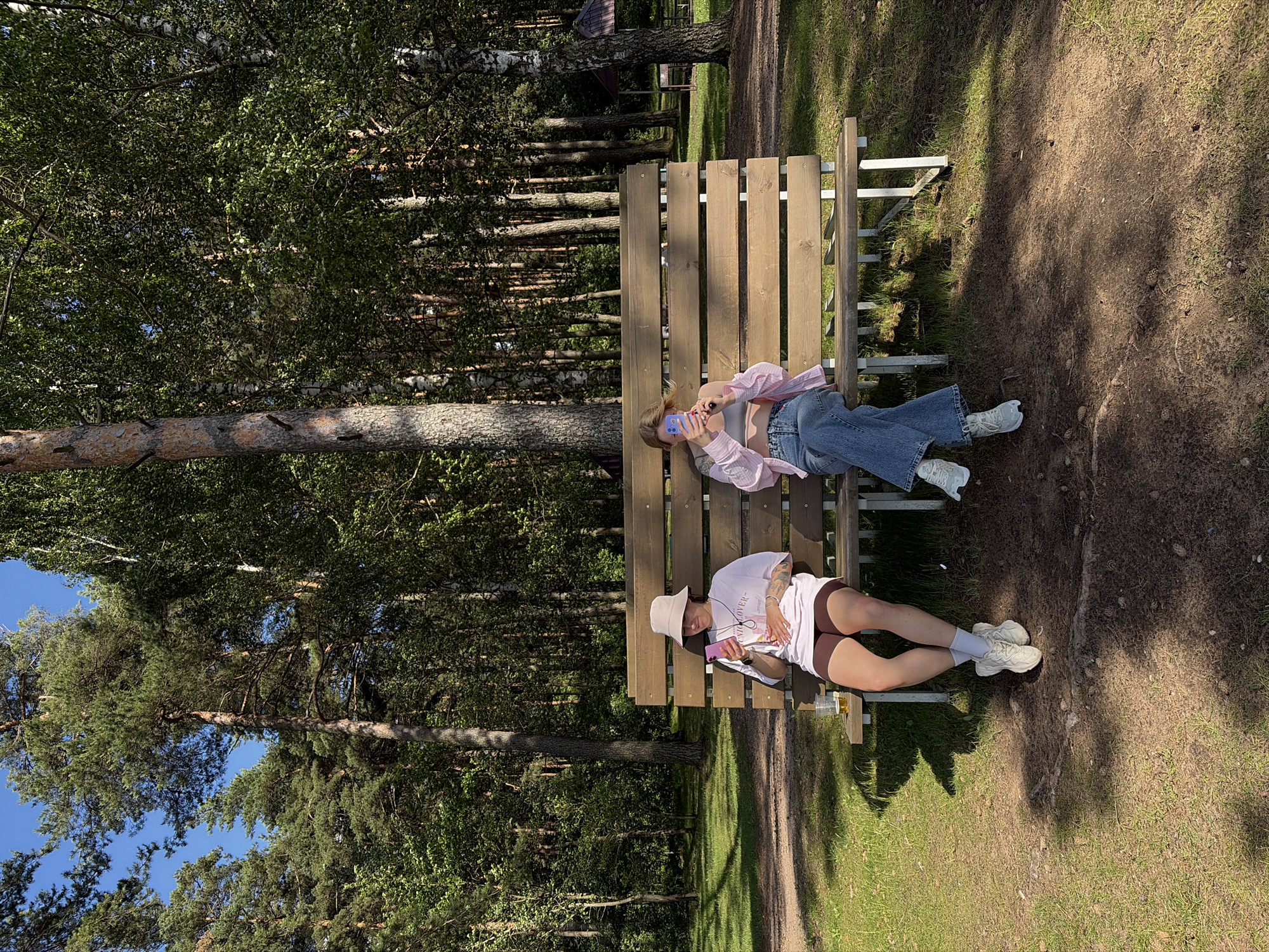 Two young women sit on a wooden park bench in a wooded area. The woman on the left wears a light-colored bucket hat, a white t-shirt, and brown shorts, holding a pink smartphone and a plastic cup. The woman on the right wears a pink open button-down shirt over a gray crop top and blue jeans, also holding a pink smartphone. They are seated at a distance from each other. The bench is situated on a dirt path with green grass and trees in the background. The environment suggests a park or recreational area within or near Ждановіцкі сельскі Савет, Belarus, characterized by dense coniferous and deciduous trees and dappled sunlight.