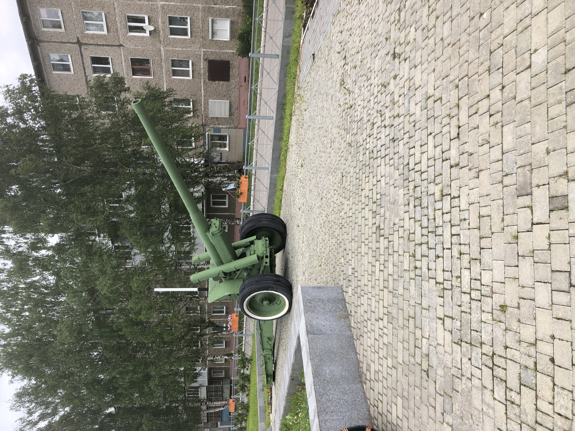 The image depicts a historical artillery piece painted green, displayed on a paved area in front of a multi-story building. The artillery piece has large wheels and a long barrel. The surrounding environment includes trees, a metal fence, and a sidewalk. The building appears to be an apartment complex with several windows. The scene is set in Solikamsk, Russia.