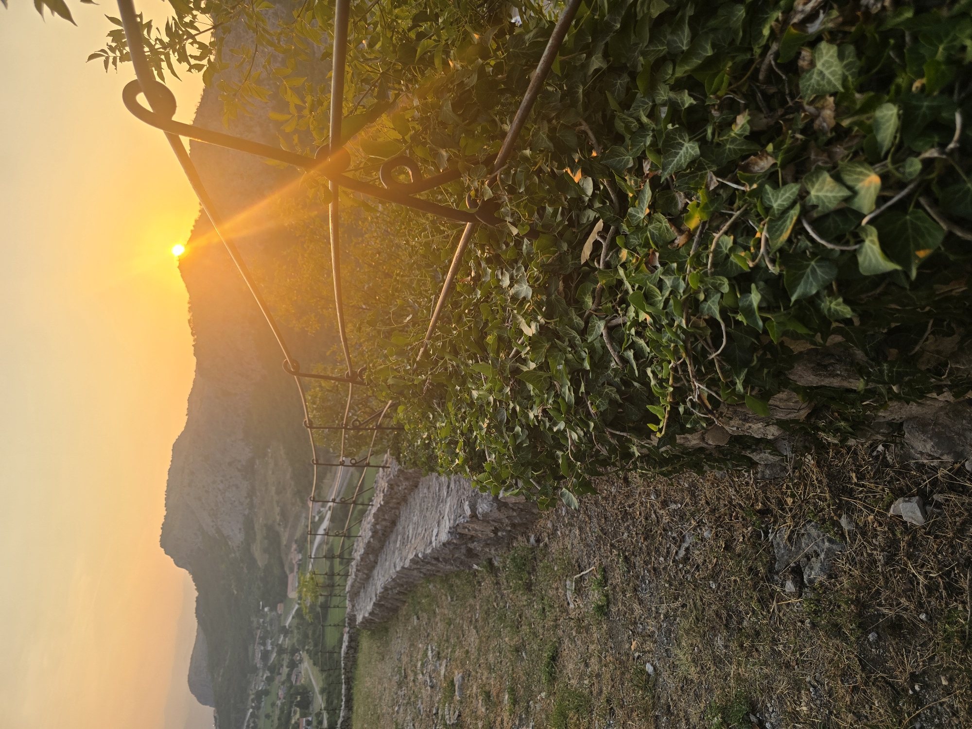 This photograph captures a sunset scene in the vicinity of Gornja Vrbica, Montenegro. The foreground is dominated by a winding stone wall covered in lush green ivy, with a gravel and dry grass path to its left. A metal railing runs along the top of the wall, separating it from the landscape beyond. In the mid-ground, a stone walkway curves along the wall, and further back, a town with scattered houses is visible nestled in a valley. The background is characterized by rolling hills and mountains, bathed in the warm, golden light of the setting sun. The sun itself is visible, peeking over the horizon of a distant mountain, casting long rays of light across the scene and creating lens flares. The sky is a vibrant gradient of orange and yellow. There are no people or animals present in the image. The overall atmosphere is serene and picturesque.