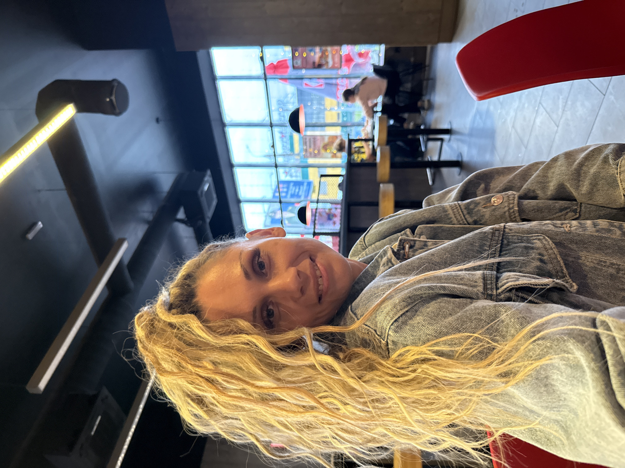 A woman with long, blonde, curly hair pulled back in a ponytail is seated in what appears to be a cafe or restaurant, based on the presence of tables, chairs, and patrons visible through a large window. She is wearing a denim jacket and looking at the camera with a smile. The interior of the establishment features dark ceilings with modern lighting fixtures and some exposed ductwork. Outside the window, city lights and signs are visible, suggesting an urban setting. The flooring is tiled, and a bright red chair is prominent in the foreground. The overall impression is a casual, indoor portrait with a blurred background.