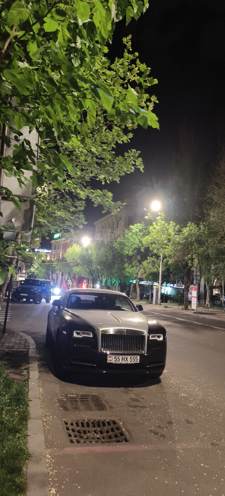 The image depicts a black and silver Rolls Royce parked on the side of a street at night. The car has an Armenian license plate that reads "55 MX 555". There is a blue Jeep visible in the background, along with street lamps and trees lining the road. A building is visible in the distance. The foreground shows a storm drain on the street next to the curb where the car is parked. The location is in the city of Unknown, Unknown.