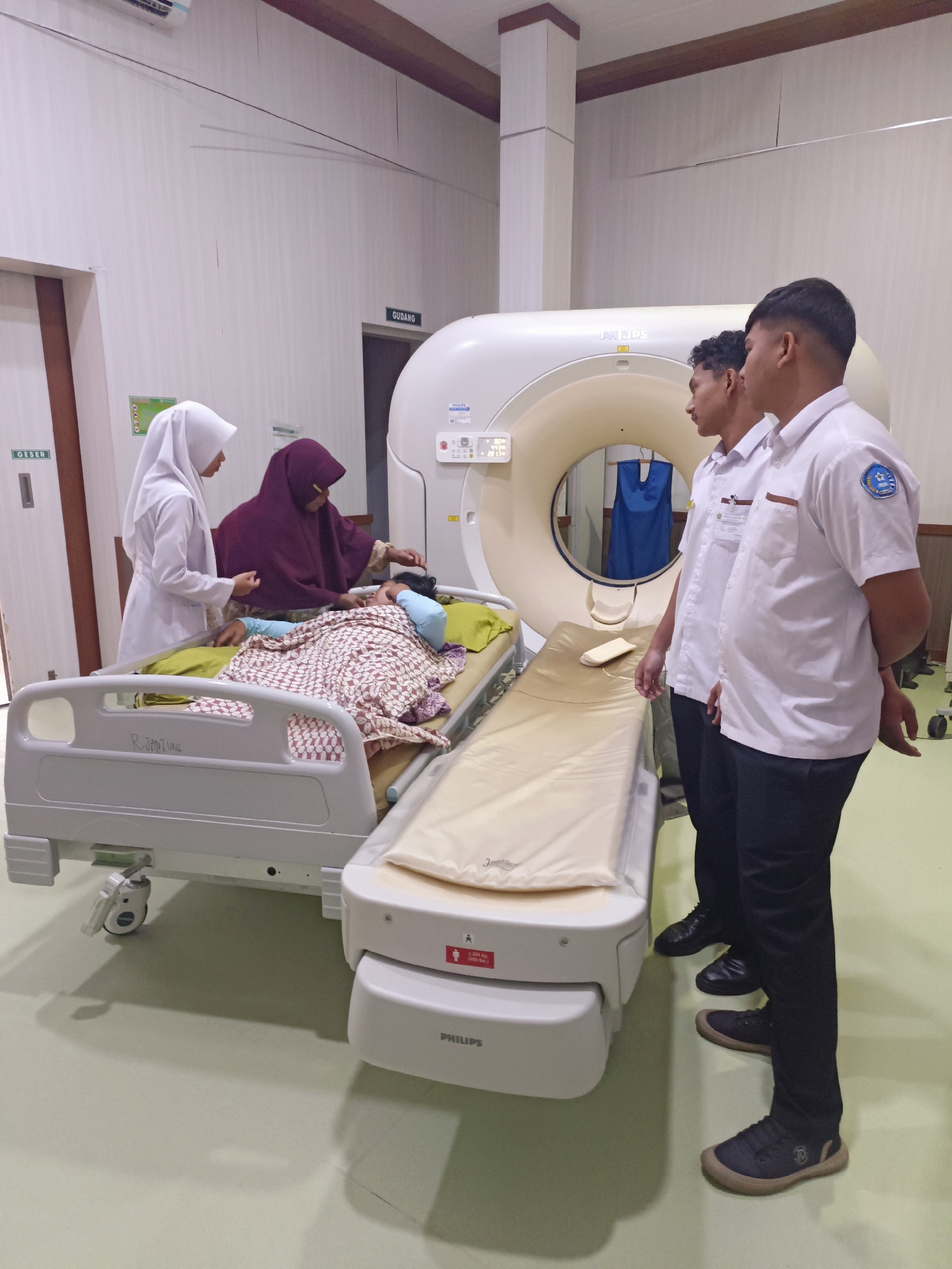 In Mesjid Reuntoh, Indonesia, a scene unfolds within a medical imaging facility, likely a hospital or clinic. Two young men, dressed in white short-sleeved uniforms with identification badges and dark pants, stand to the right, observing. In the center, a woman in a maroon hijab is attending to a patient lying on a hospital bed. The patient, covered with a patterned blanket, appears to be resting. To the left of the patient's bed, a woman in a white hijab and a white coat, presumably a medical professional, also attends to the patient. Adjacent to the patient's bed is another medical bed, unoccupied. Dominating the background is a large, white CT scanner, a prominent piece of medical equipment. The environment is a brightly lit room with pale walls and a light-colored floor. Signage on the wall indicates "GUDANG" (Storage) and "GESER" (Slide), suggesting the presence of storage areas or sliding doors. The overall impression is one of a medical procedure or training session taking place in a clinical setting.