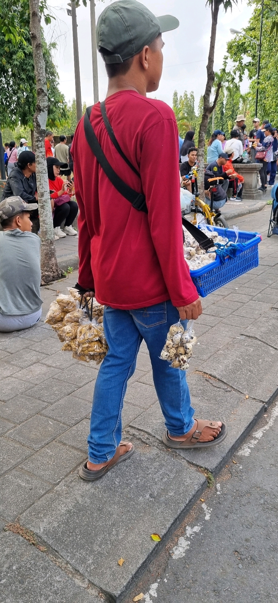 A young male, wearing a red long-sleeved shirt, blue jeans, a green baseball cap, and brown sandals, walks on a paved sidewalk in Denpasar, Indonesia. He is carrying a blue plastic basket filled with food items on his right side, with a black strap across his body. In his left hand, he holds two bags of quail eggs, and in his right hand, he holds several bags of food items.
In the background, other people are visible, some sitting and others standing, and trees line the area. The pavement is made of interlocking grey stones. The scene appears to be outdoors, possibly in a park or a public gathering area. The overall impression is one of a typical street scene in a bustling urban environment.