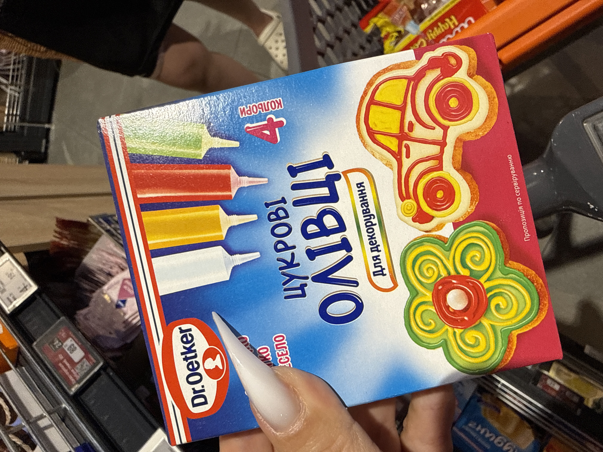 The image shows a close-up of a box of Dr. Oetker icing decorations. The box is primarily blue and red, with images of decorated cookies in the shapes of a car and a flower. The text on the box is in Ukrainian. Visible text includes "Dr. Oetker", "КОЛЬОРИ 4" (Colors 4), "ЦУКРОВІ ОЛІВЦІ" (Icing Pencils), and "Для декорування" (For decorating). There is also a smaller text line reading "Пропозиція по сервіруванню" (Serving suggestion).
The box is held by a person whose hand and fingernail are visible in the foreground. The fingernail is long and white. The background is blurred but suggests a retail environment, possibly a supermarket aisle, with shelves and other products visible. The lighting appears to be artificial, typical of an indoor store. The setting is identified as Odesa, Ukraine, though there are no specific visual cues in the image to confirm this location. The overall impression is of someone browsing or purchasing baking supplies.