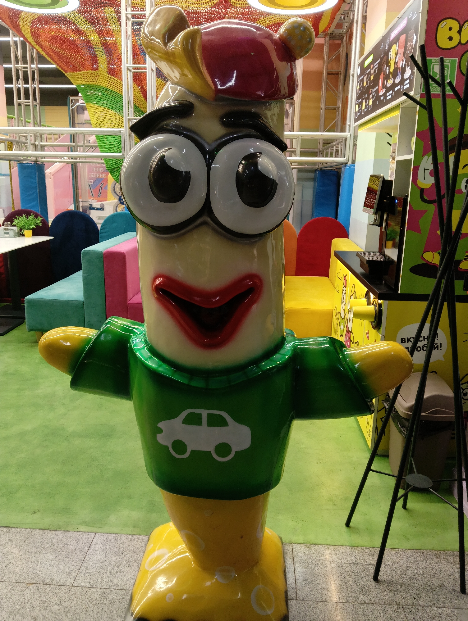 An animated, cartoon-like figure stands in what appears to be a children's play area. The figure is predominantly yellow and white with large black and white eyes, a wide red mouth, and a green shirt featuring a white car graphic. The figure has yellow arms and feet. Behind the figure, brightly colored seating in blue, green, and pink is visible. The floor is covered in a lime green carpet. In the background, there is a structure with netting in rainbow colors, and to the right, a black metal stand with thin legs holds up some black rods. A trash can is also visible. A sign with writing and illustrations is present on the right side, and a digital screen is mounted on a black surface. The environment suggests a commercial or entertainment venue, possibly a cafe or a play center. The image is a medium shot, with the figure taking up most of the foreground. The specific location in Hrodna, Belarus, is not discernible from the visual information provided in the image.