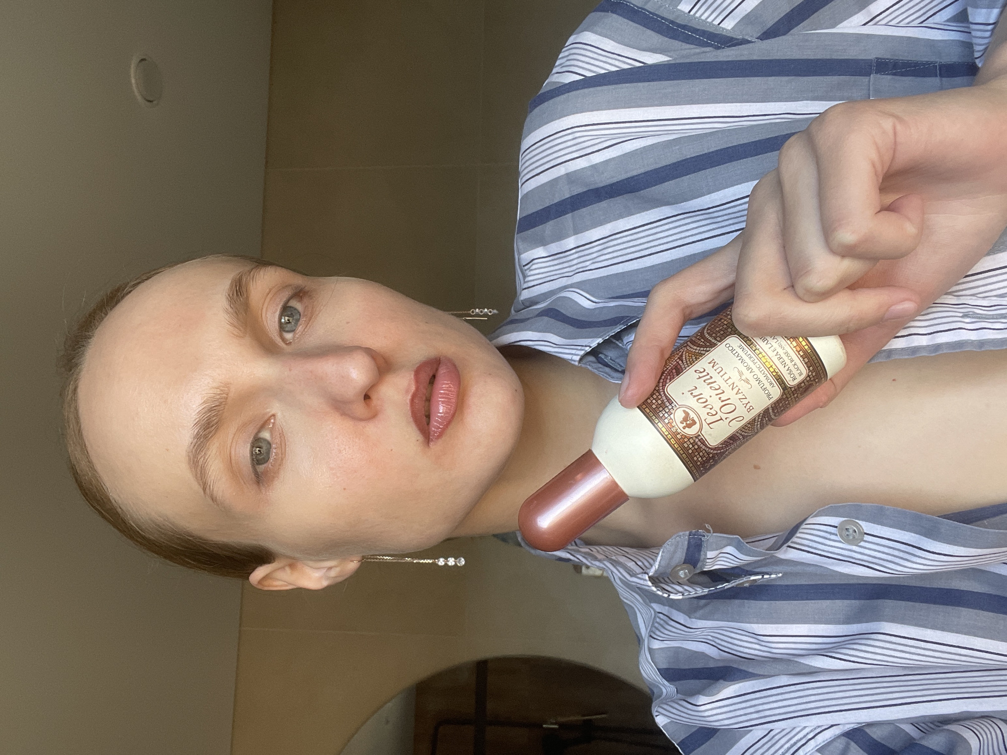 A person is holding a bottle of Tesori d'Oriente Byzantuim perfume. The person has fair skin and is wearing a striped blue and white button-up shirt. Their hair is light brown and pulled back. They are wearing a long, dangly earring. The bottle has a cream-colored label with brown details and a rose gold cap. The background is a plain light brown wall.