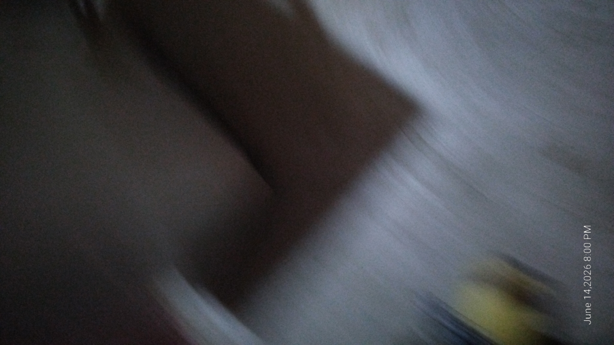 The image is a blurry, low-light photograph with a date and time stamp of "June 14, 2026 8:00 PM" in the bottom left corner. The scene appears to be indoors, possibly a bedroom, with soft, diffused lighting. The dominant colors are muted grays, blacks, and browns, with a hint of yellow in the lower left quadrant. The lack of sharp focus and detail makes it difficult to identify specific objects or subjects. The image does not contain any discernible features that would allow for the determination of a specific location within the city of Howayi, Nigeria.