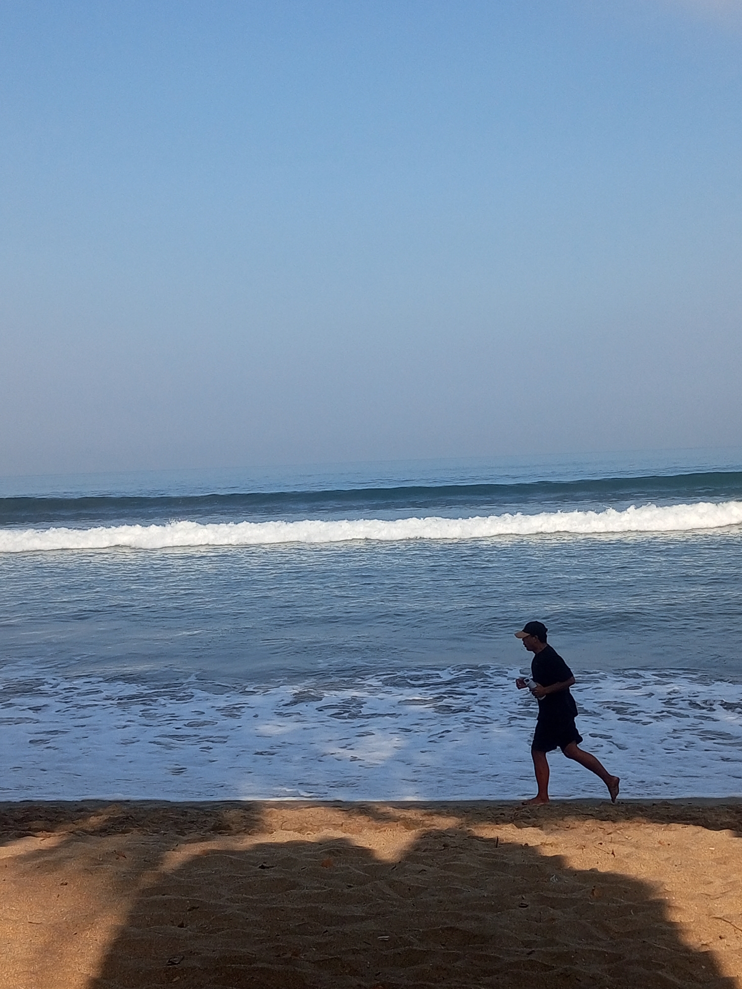 A man in a black t-shirt and shorts, wearing a baseball cap, jogs along the sandy shore of Kuta Beach, Bali, Indonesia. The ocean waves crash onto the beach, and the sky is a clear, soft blue. The man appears to be holding a bottle in his right hand. Shadows of palm trees or other foliage are cast on the sand in the foreground.