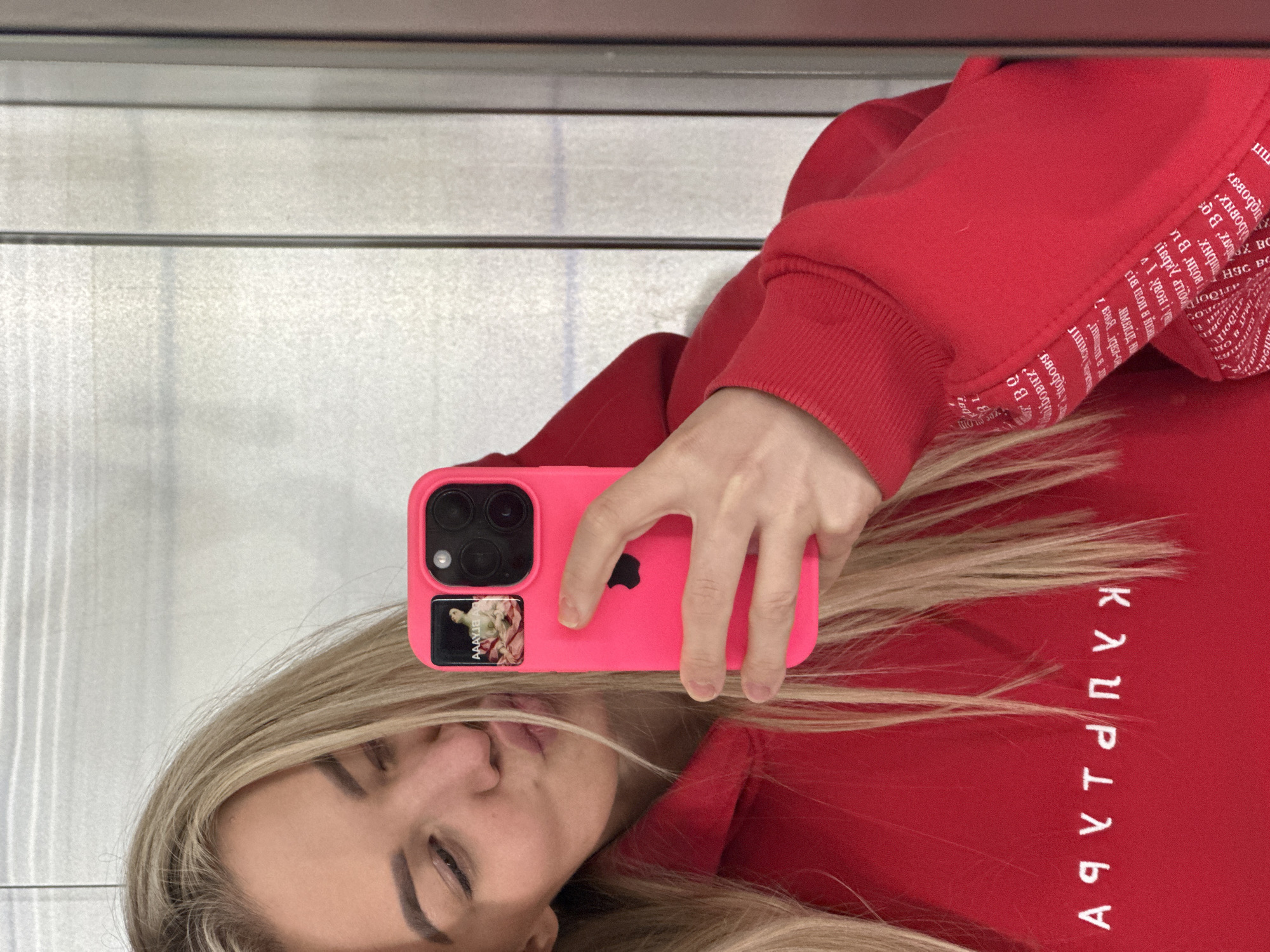 An overhead shot depicts a person taking a selfie. The subject is a woman with long blonde hair, seen from the chest up, with her head tilted. She holds a bright pink smartphone with her right hand, positioned above her head. Her eyes are mostly closed or looking down, and her lips are slightly parted. She is wearing a red hooded sweatshirt.
The smartphone is an iPhone model with three cameras. The pink phone case has a small rectangular sticker below the camera module, depicting a classical painting, possibly of a woman, with the visible text "AAAYJE". The red sweatshirt features white Cyrillic text printed repeatedly across the fabric, particularly noticeable on the sleeves and the back. Words like "любов" (love) and "свобода" (freedom) are discernible on the sleeve, while the word "КАРТИНА" (Kartina, meaning painting) is visible and mirrored on the back of the sweatshirt due to the reflective nature of the image.
The background appears to be a light-colored, possibly textured wall or surface, with horizontal lines. The image seems to be a reflection, likely taken in a mirror or similar reflective surface. There are no visual cues within the image to suggest the provided location context of Budva, Montenegro.