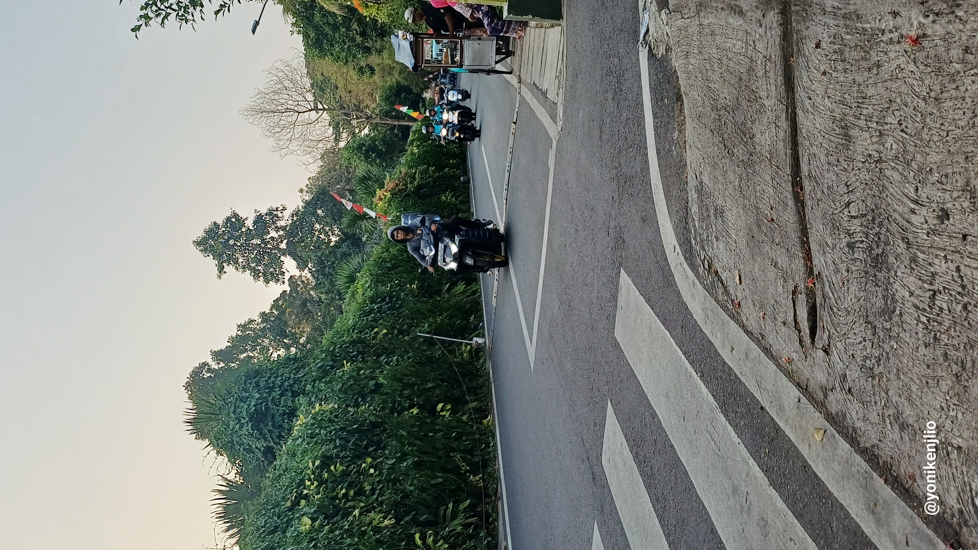 In a moderately lit outdoor scene, a person wearing a grey hooded jacket rides a black motorcycle forward on a paved road. The motorcycle has its headlights on. Behind this motorcycle, two more motorcycles are visible, also traveling in the same direction. To the right of the road, a small food cart with a blue awning is parked, and three people are standing near it, two of whom appear to be customers and one vendor. The road is marked with white pedestrian crossing lines in the foreground. Lush green foliage lines the left side of the road, creating a dense hedge. The sky above is overcast with a light, hazy appearance. The overall impression is a street scene in a tropical or subtropical environment. The image also contains a watermark with the text "@yonikenjilio" at the bottom left corner, suggesting the image's origin.
