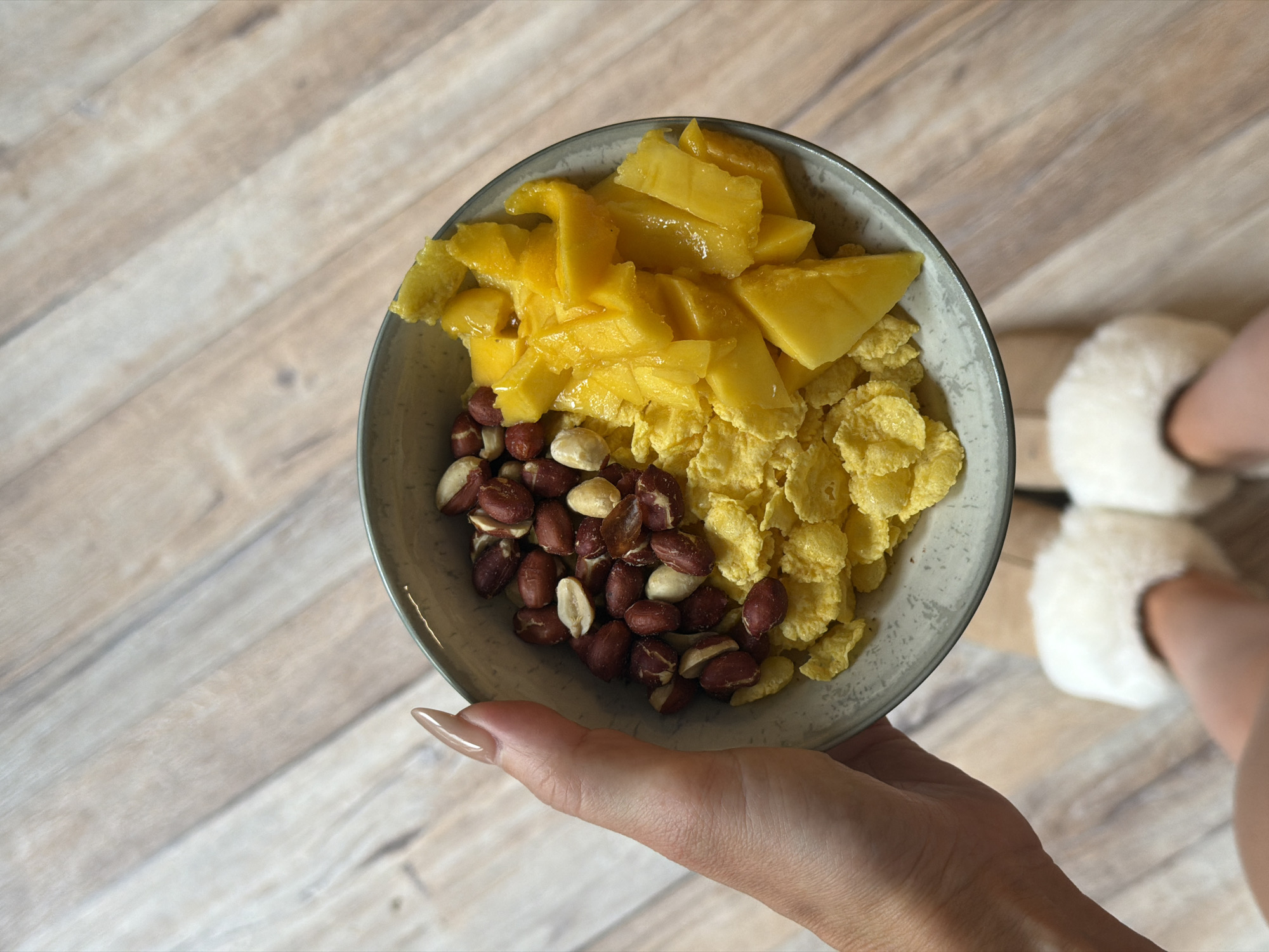 A close-up overhead shot shows a light grey bowl filled with a breakfast assortment. The bowl is held in a person's hand, which is visible in the upper left corner of the frame. The bowl contains golden corn flakes topped with chopped mango pieces and a mix of peanuts. The background consists of a light-colored wooden floor. The image is positioned in Perm, Russia.